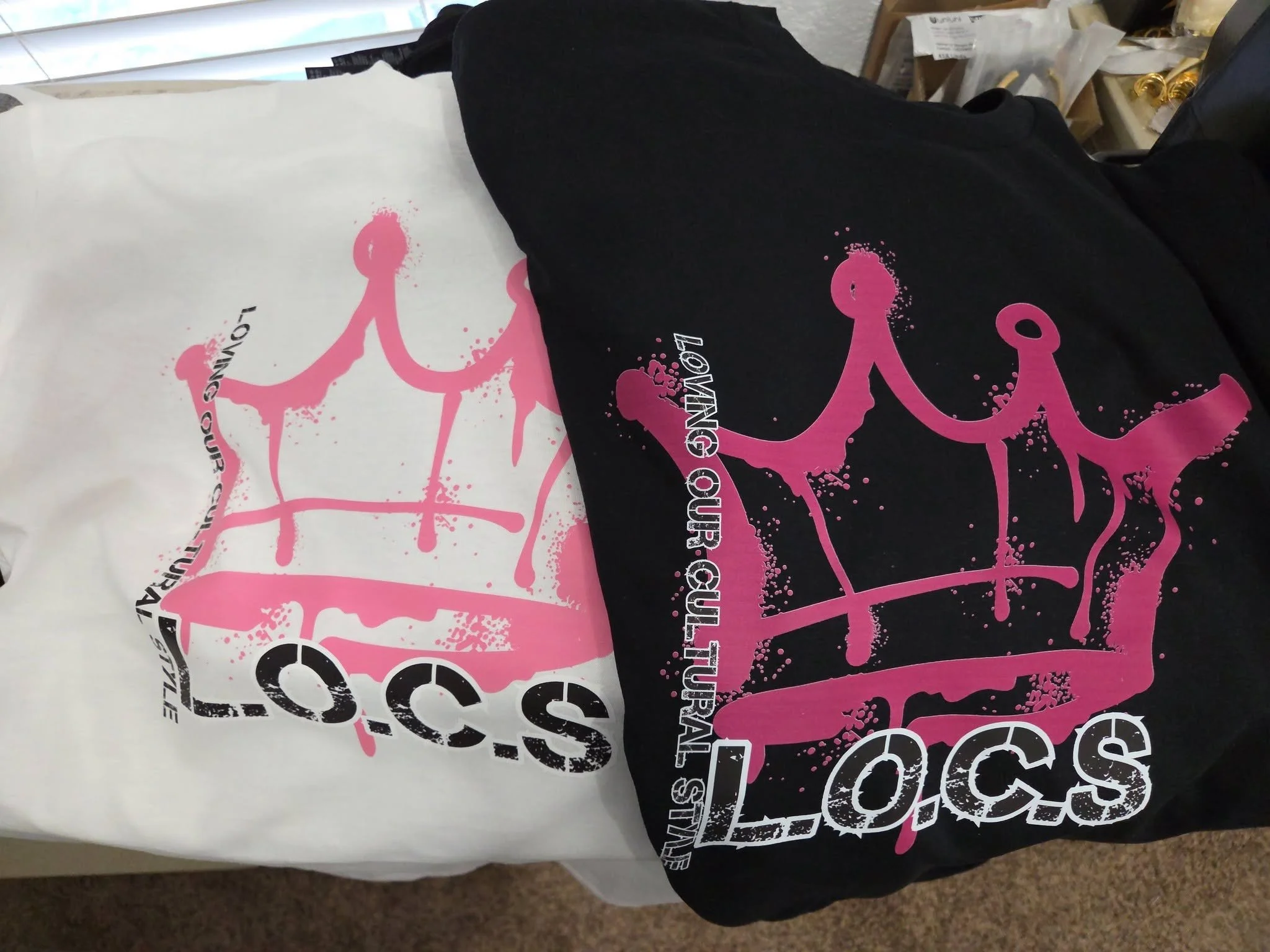 Close-up view of two t-shirts, one black and one white, with pink and black graphic designs and text, lying on a flat surface.