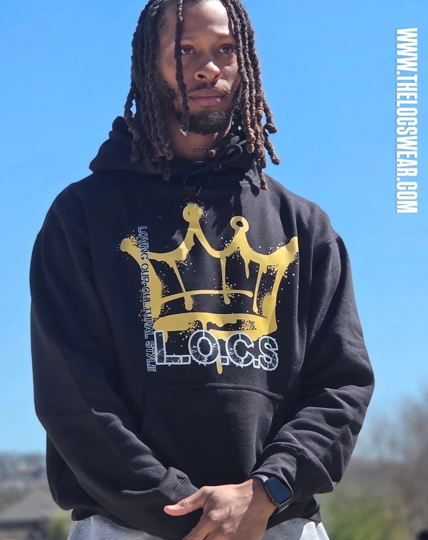 A man with long locs and a beard standing outdoors with a clear blue sky in the background, wearing a black hoodie with a gold crown and text design, and a smartwatch on his left wrist.