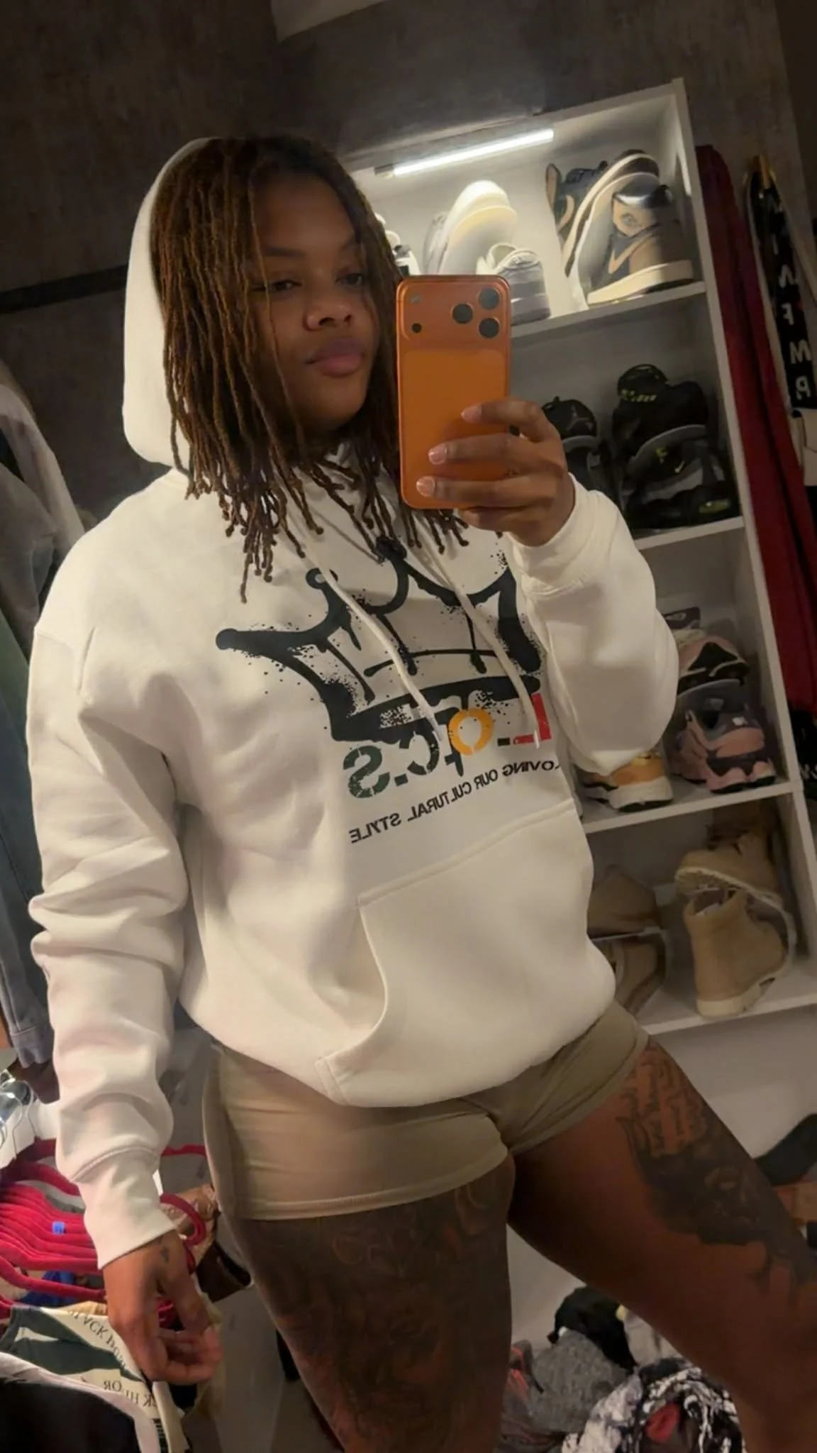 A woman with shoulder length locs taking a mirror selfie in her closet, wearing a white hoodie that has a black 'LOCS' crown and shorts, with a tattoo on her thigh. Shelves in the background display shoes and sneakers.