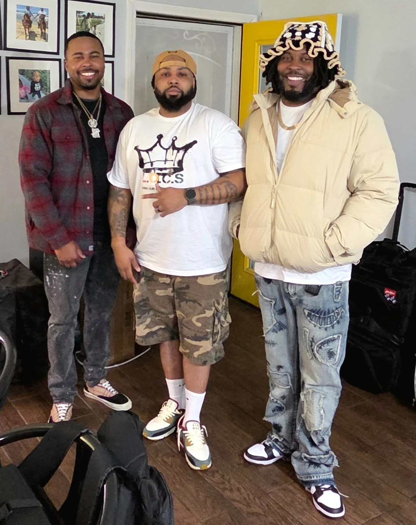 Group of three men standing together indoors, smiling at the camera. One is wearing a red plaid shirt, the middle man is in a white T-shirt and camouflage shorts, and the third is in a beige jacket with a patterned hat and distressed jeans.