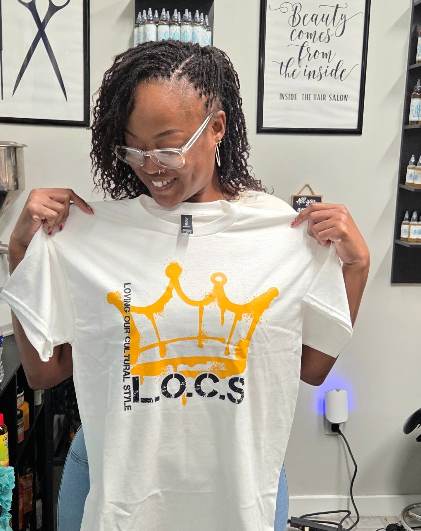 Woman smiling and holding a white T-shirt with a yellow crown graphic and the text 'L.O.C.S' and 'Loving Our Cultural Style' printed on it, inside a hair salon with framed signs on the wall.