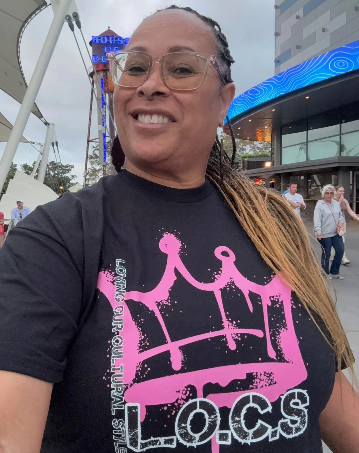 A smiling person wearing glasses and a black T-shirt with pink graffiti-style crown and the word 'LOCS Loving our Cultural Style' text, standing outdoors near a Ferris wheel and a modern building, with other people in the background.