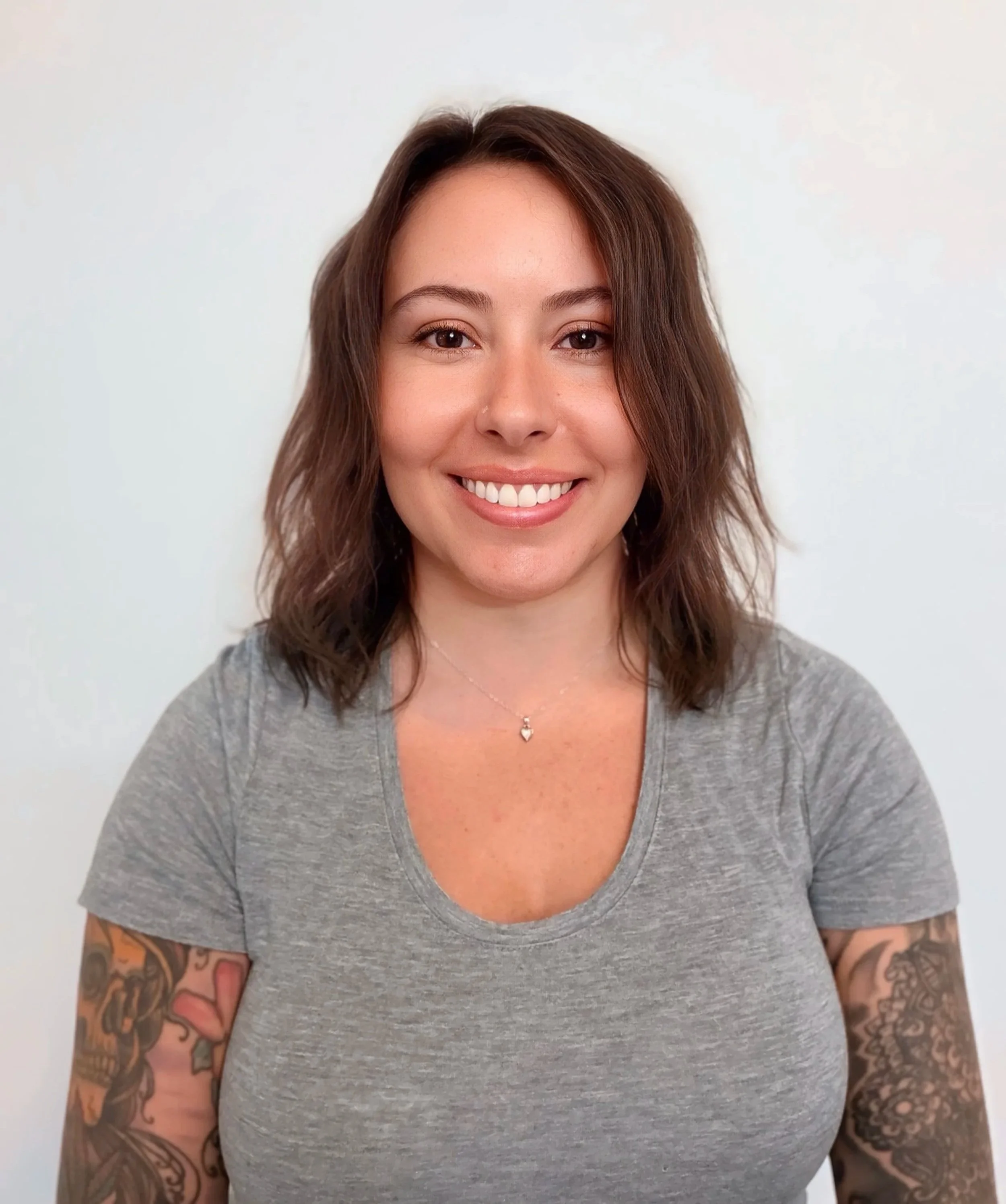 A woman with shoulder-length brown hair, smiling, wearing a gray V-neck T-shirt, with tattoos on both arms, in front of a plain white background.