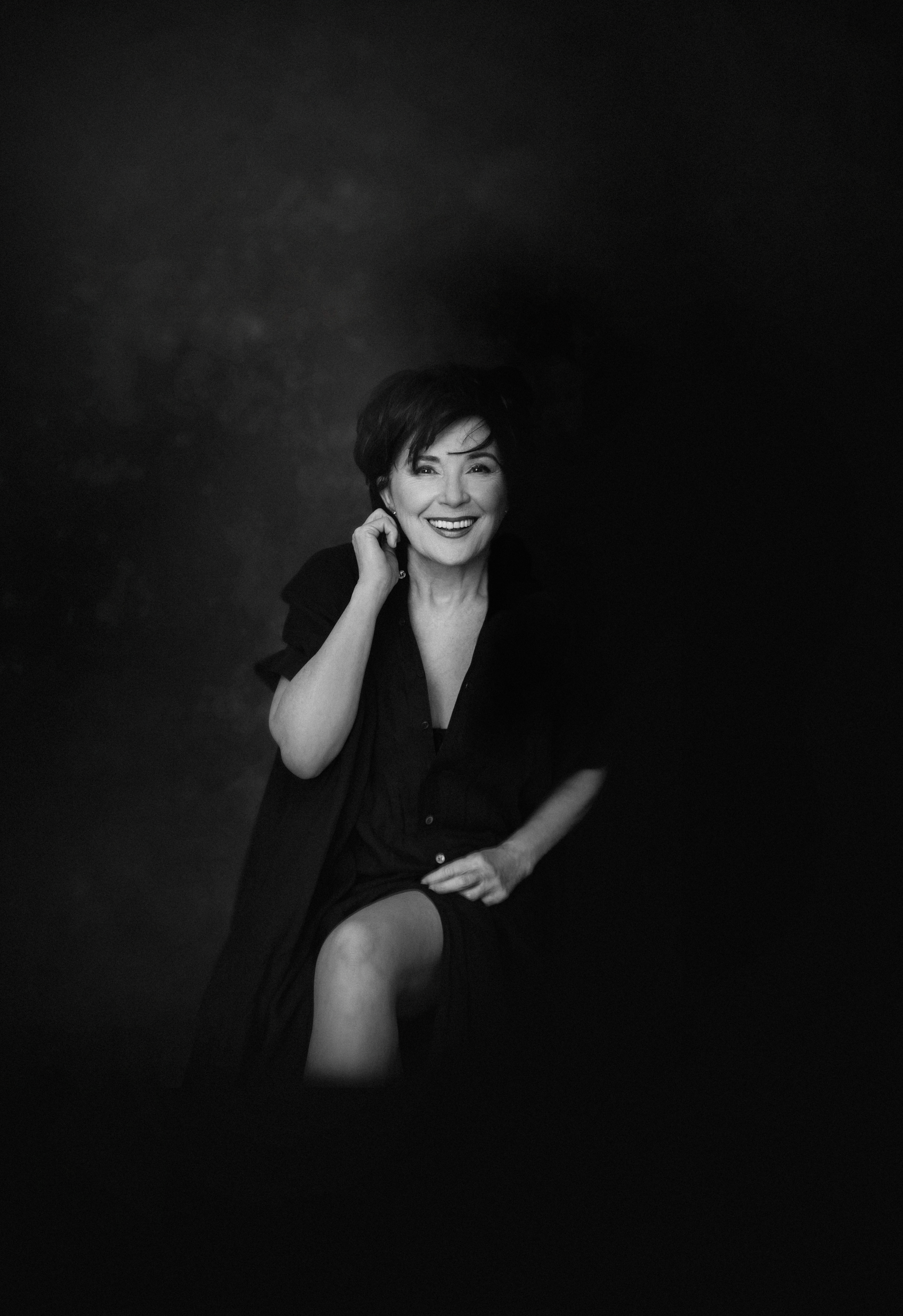 Une femme souriante en noir et blanc, portant une robe noire, assise et levant une jambe, dans un décor sombre.