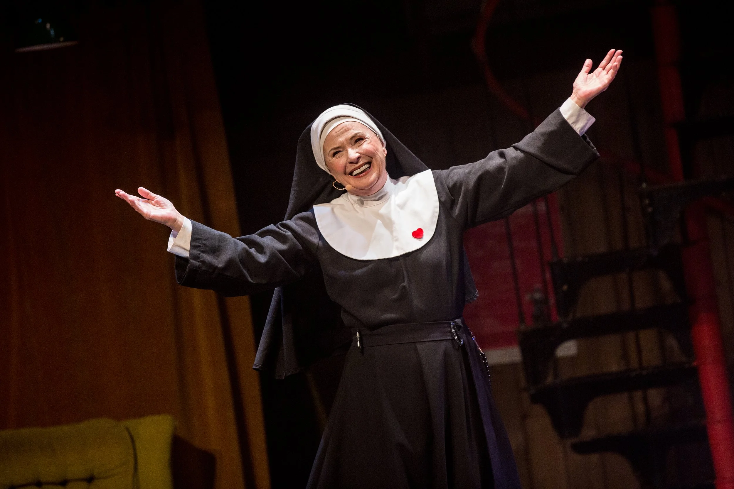 Une femme habillée en religieuse avec une grande tendance joyeuse, bras écartés, en train de s'exprimer sur scène dans un théâtre avec un fond sombre.