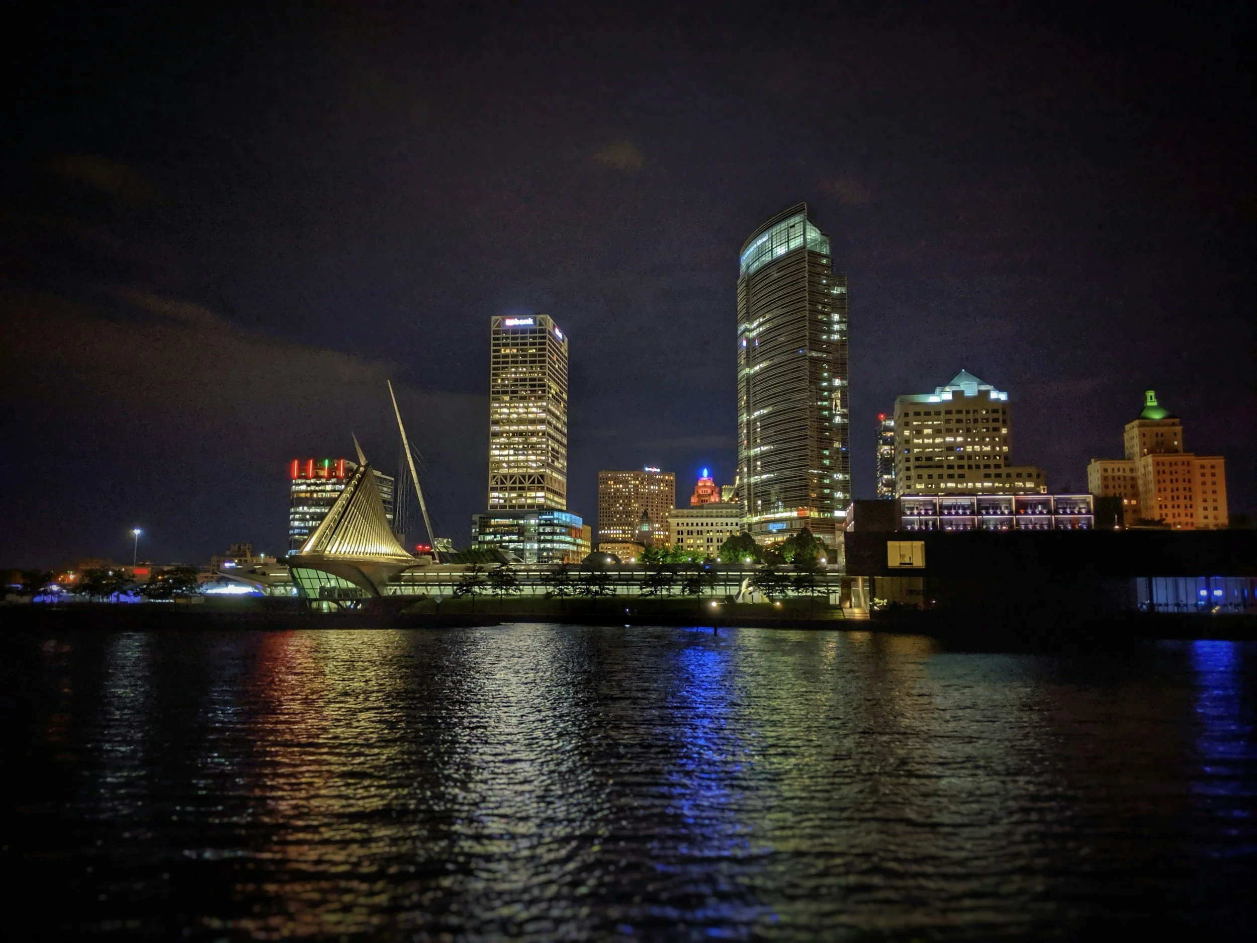 Nighttime city skyline with tall buildings illuminated, water in the foreground reflecting city lights, and a dark sky above.