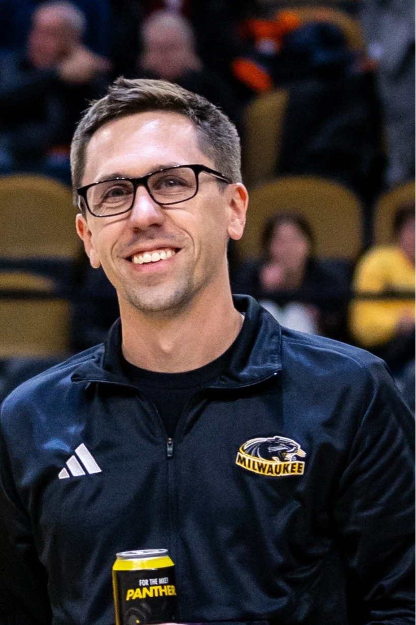 A smiling man wearing glasses and a black Adidas jacket with Milwaukee Panthers logo, holding a can.