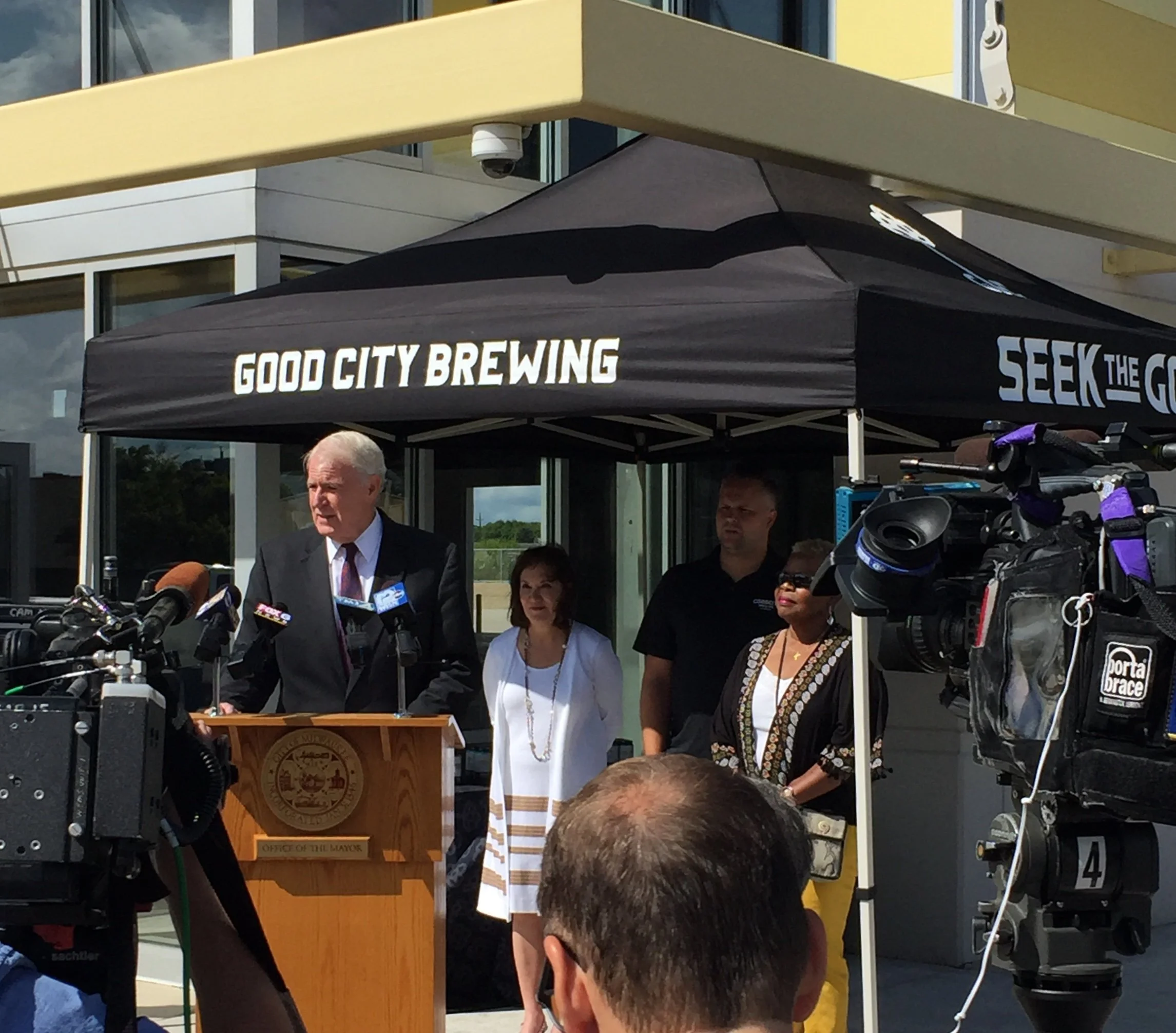 A speaker at a podium labeled 'Office of the Mayor' speaking at an outdoor event, with a canopy with 'Good City Brewing' written on it. Several people, including men and women, are gathered around, and there are cameras and microphones set up.