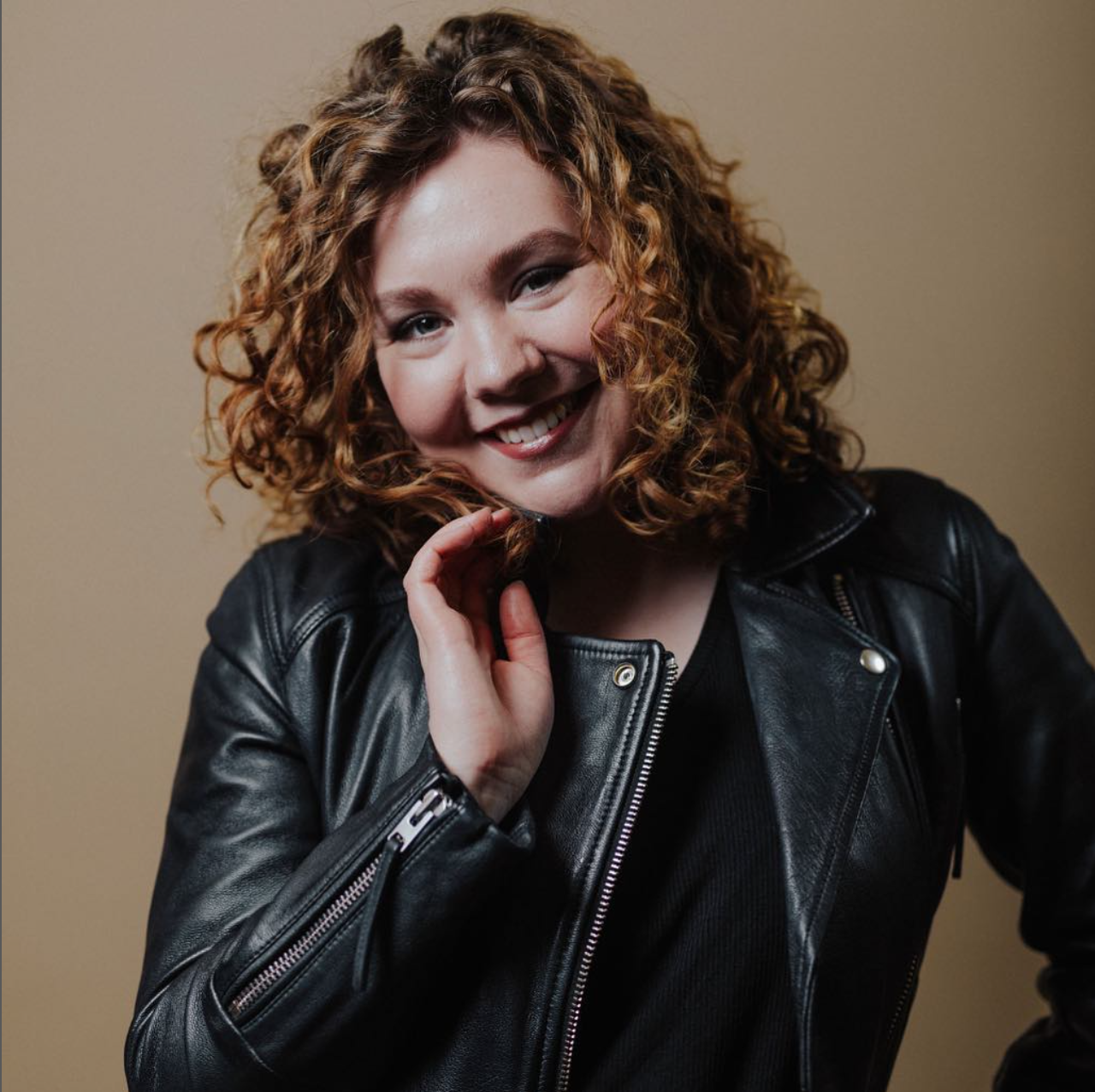 Portrait of a woman with curly red hair smiling, wearing a black leather jacket, against a beige background.