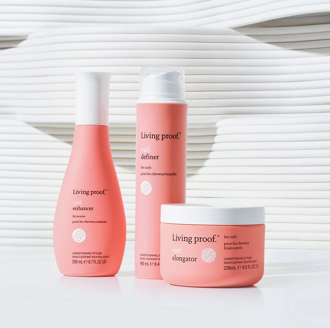 Three pink hair care bottles from Living Proof, including a curl enhancer, curl definer, and curl elongator, on a white surface with a textured white background.