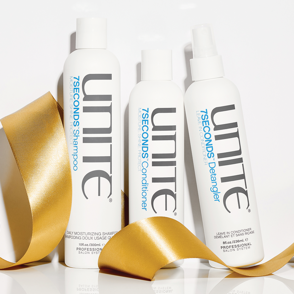 Three white bottles of UNITE hair care products on a white surface with gold ribbon decoration. The bottles are labeled daily moisturizing shampoo, conditioner, and leave-in conditioner.