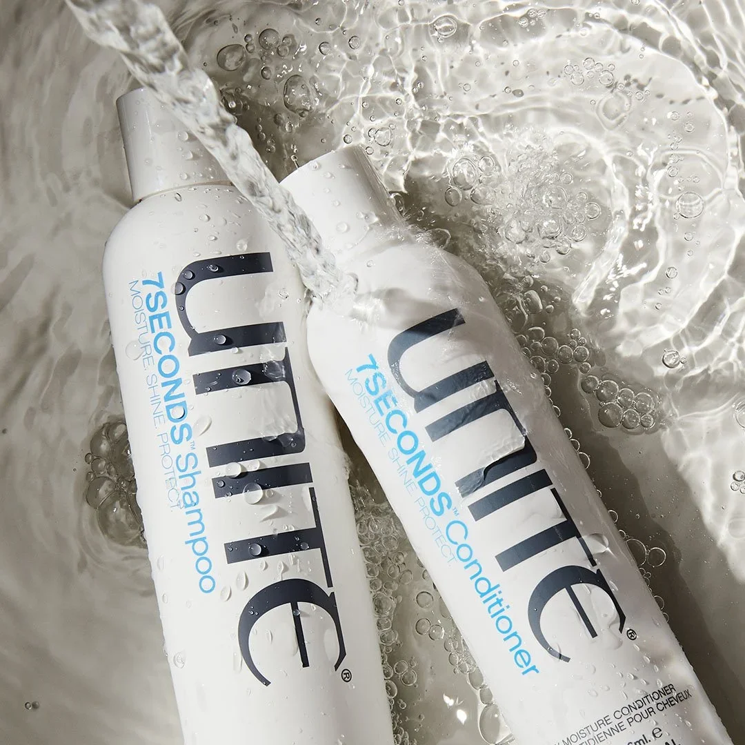 Two white bottles of UNITE shampoo and conditioner with black and blue text, under running water with bubbles.