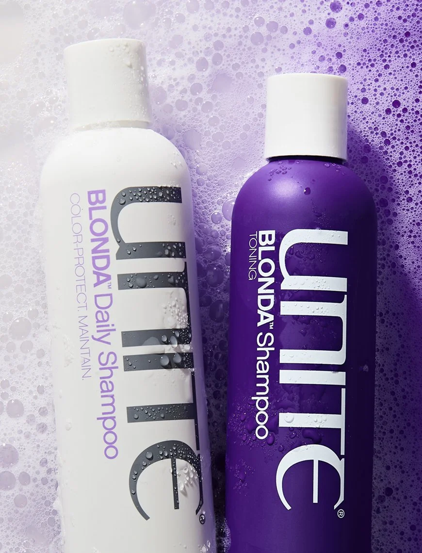 Two bottles of UNITE BLONDA toning shampoo, one white and one purple, surrounded by purple soap bubbles.