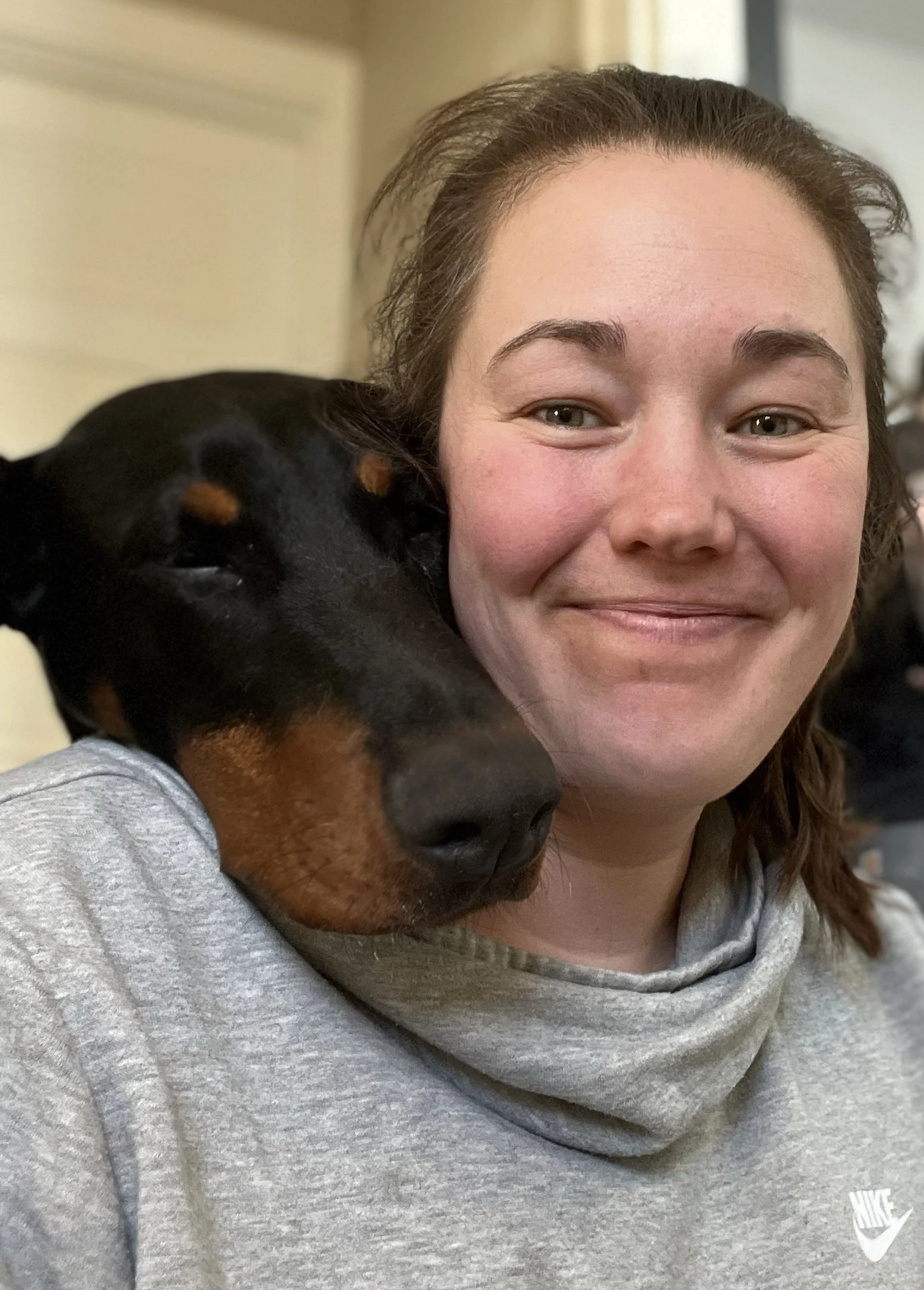 A woman smiling with a large doberman black and tan dog resting its head on her shoulder.