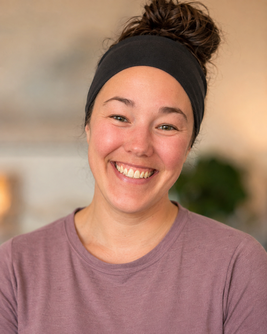 A woman with fair skin, brown hair in a bun, wearing a black headband and a mauve T-shirt, smiling broadly with visible teeth, in an indoor setting.