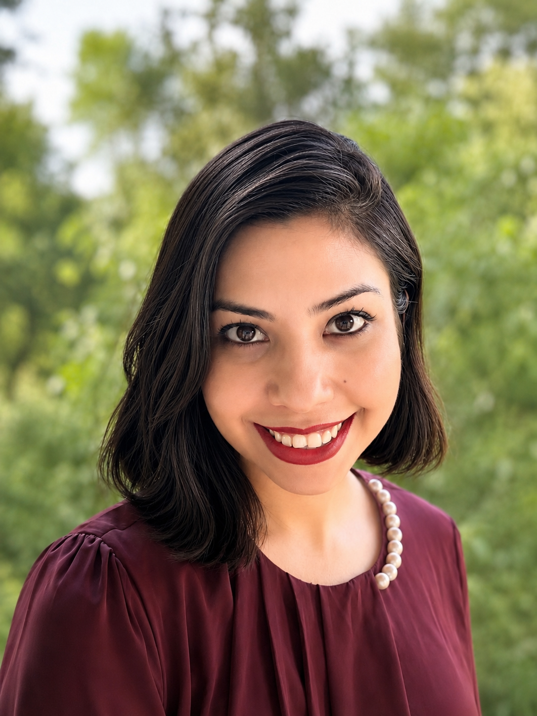 Nishat Farzana, an Academic and Researcher based in Italy. She is a woman with dark hair, smiling, wearing a maroon top and a pearl necklace, outdoors with a background of green trees.