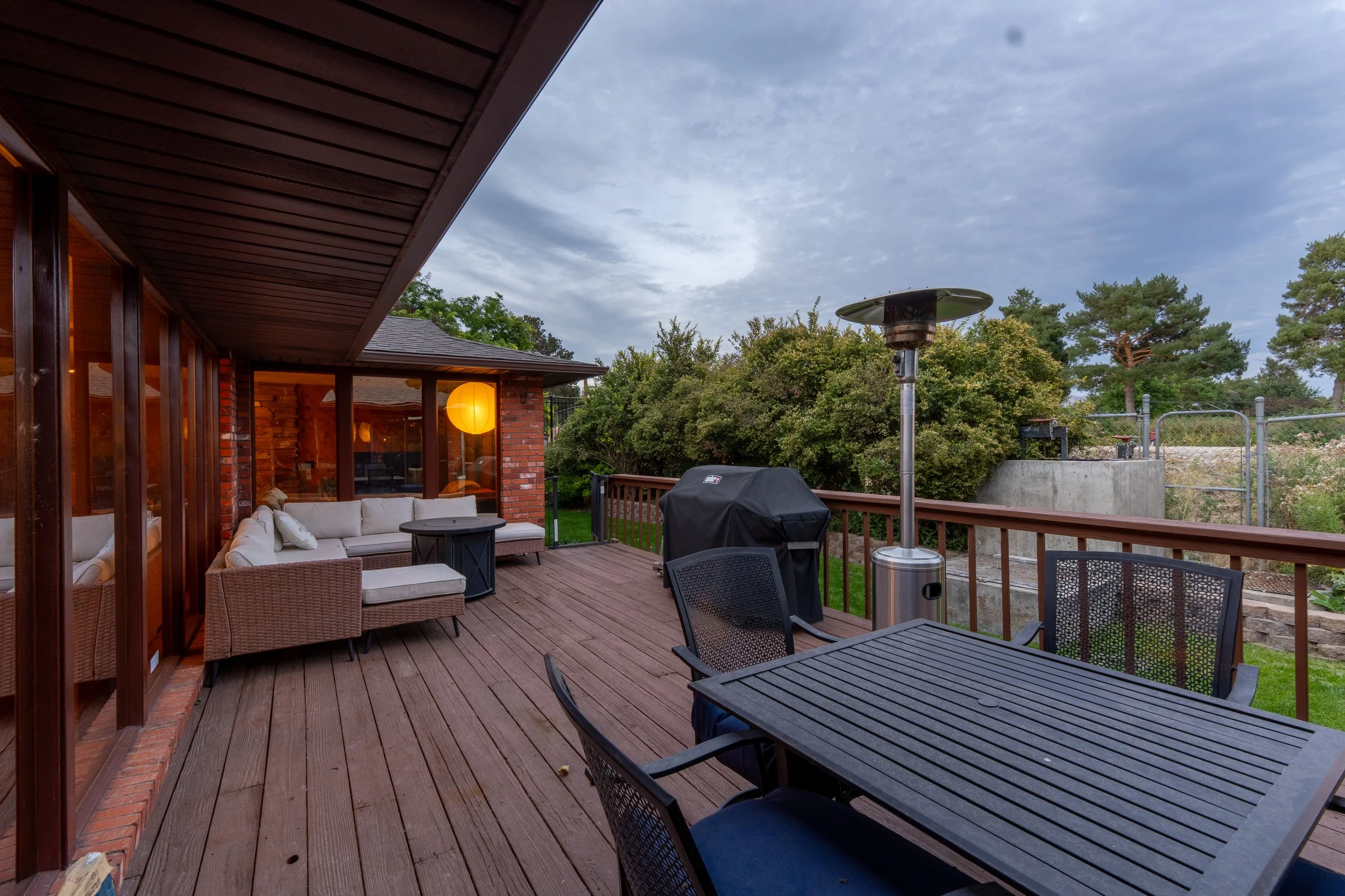 A wooden outdoor deck attached to a house with large windows and a patio area with seating, a grill, and outdoor heating, surrounded by trees and a partly cloudy sky.