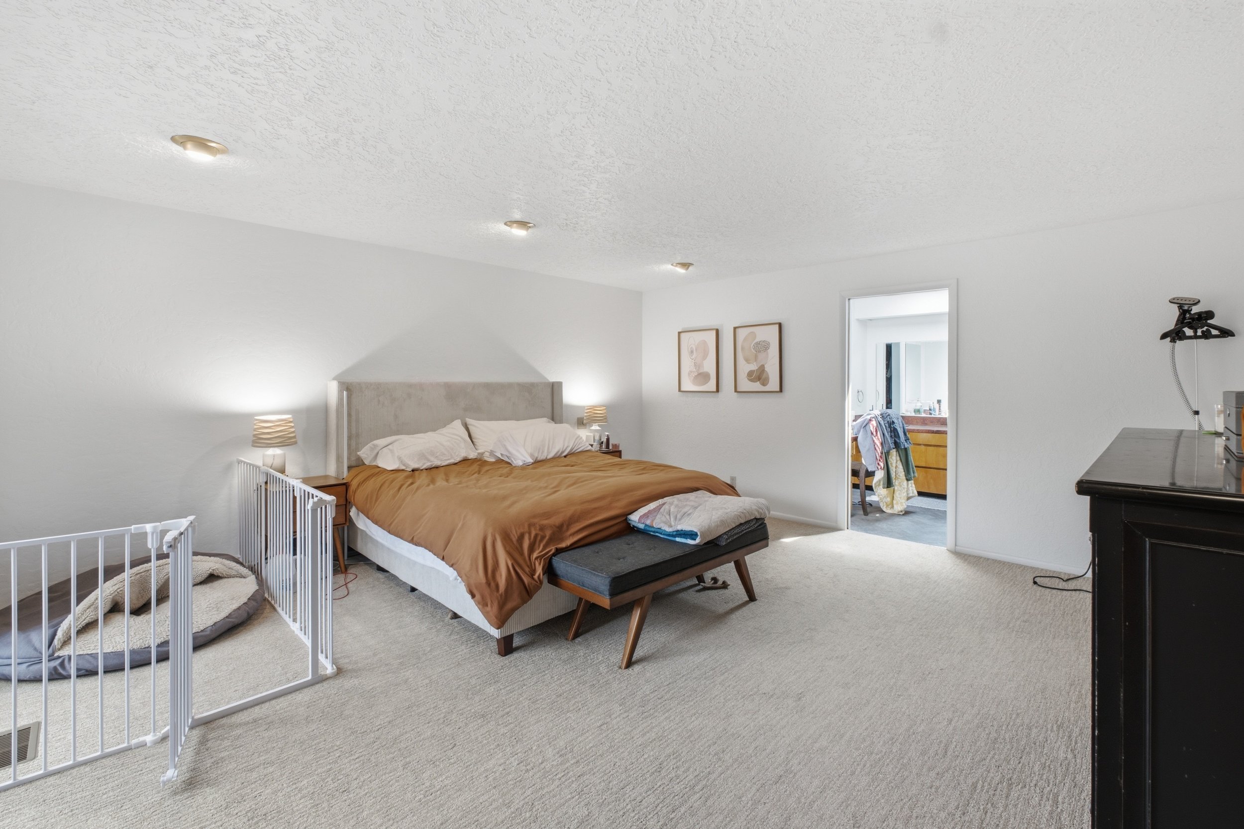 A bedroom with a bed, nightstands, lamps, wall art, and an open door leading to another room with laundry and a countertop.