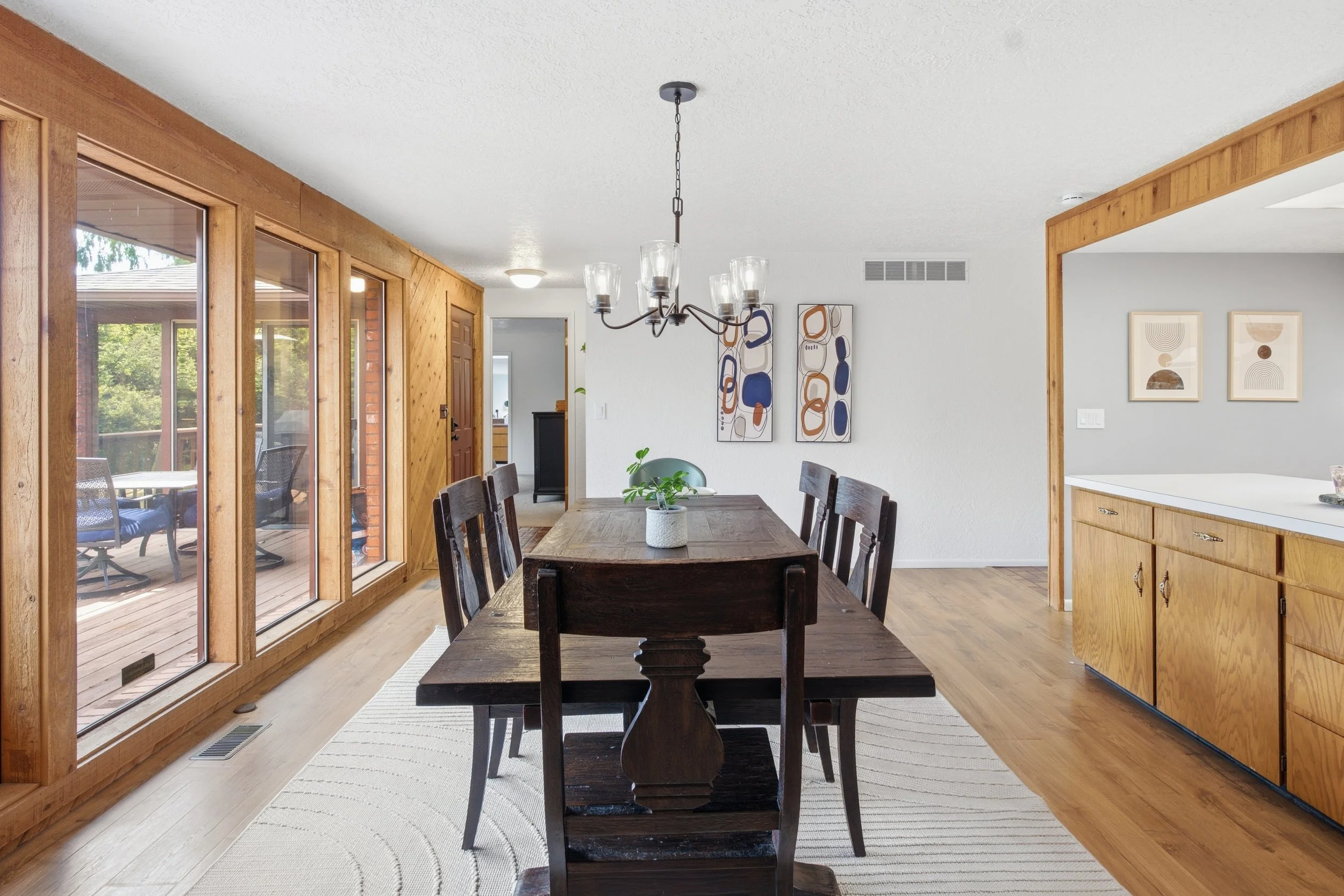 Dining room with wooden dining table and chairs, abstract wall art, large windows, and a chandelier hanging from the ceiling.