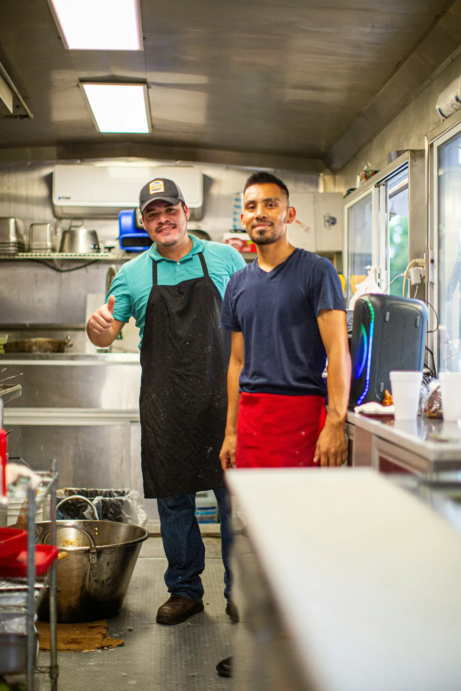 Dos hombres en un camión de comida, uno con delantal y gorra, haciendo señal de aprobación, y el otro con camiseta azul y delantal rojo, ambos sonriendo.