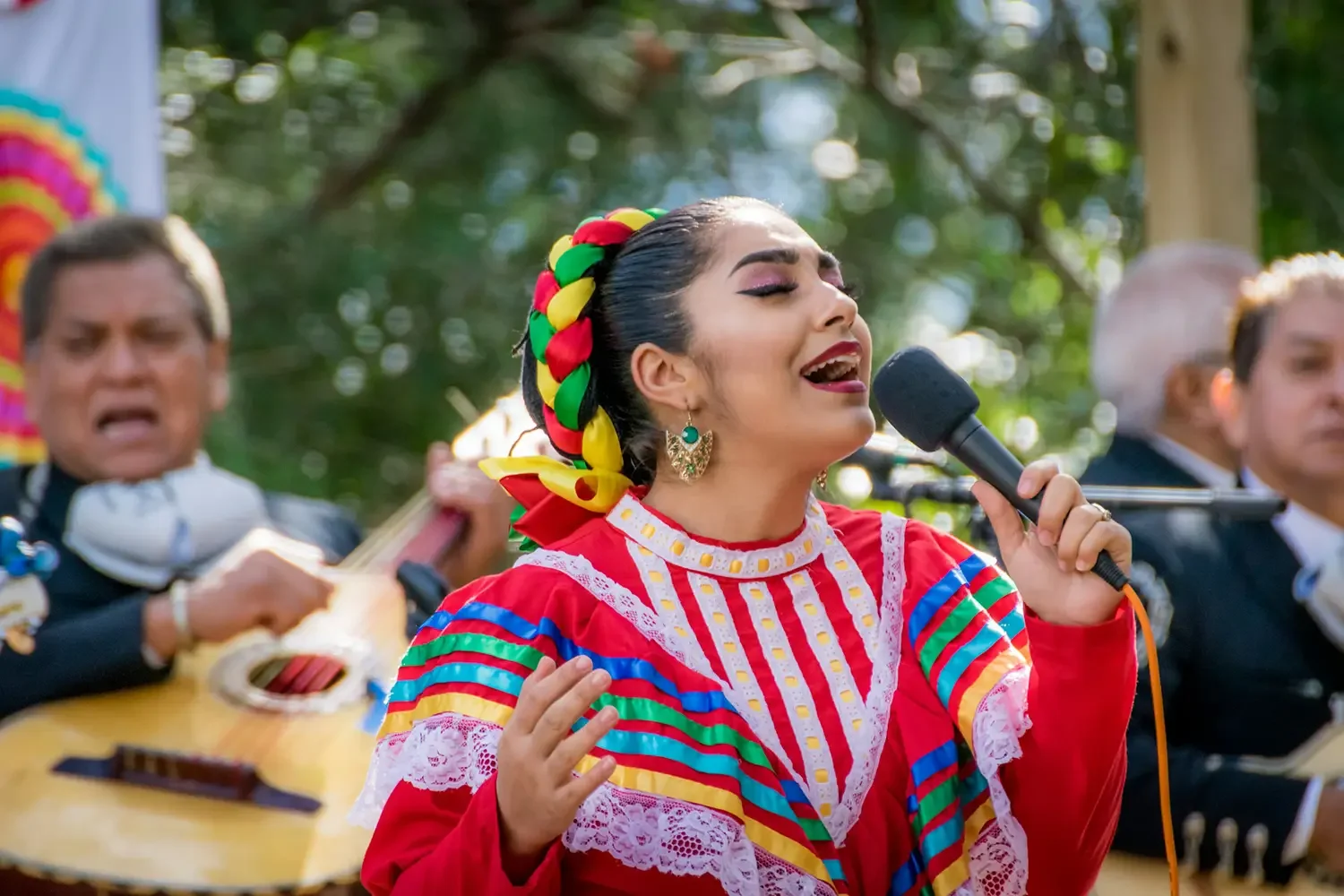 Mujer vestida con traje colorido tradicional mexicana cantando con un micrófono, acompañada por músicos con guitarras en un ambiente al aire libre.