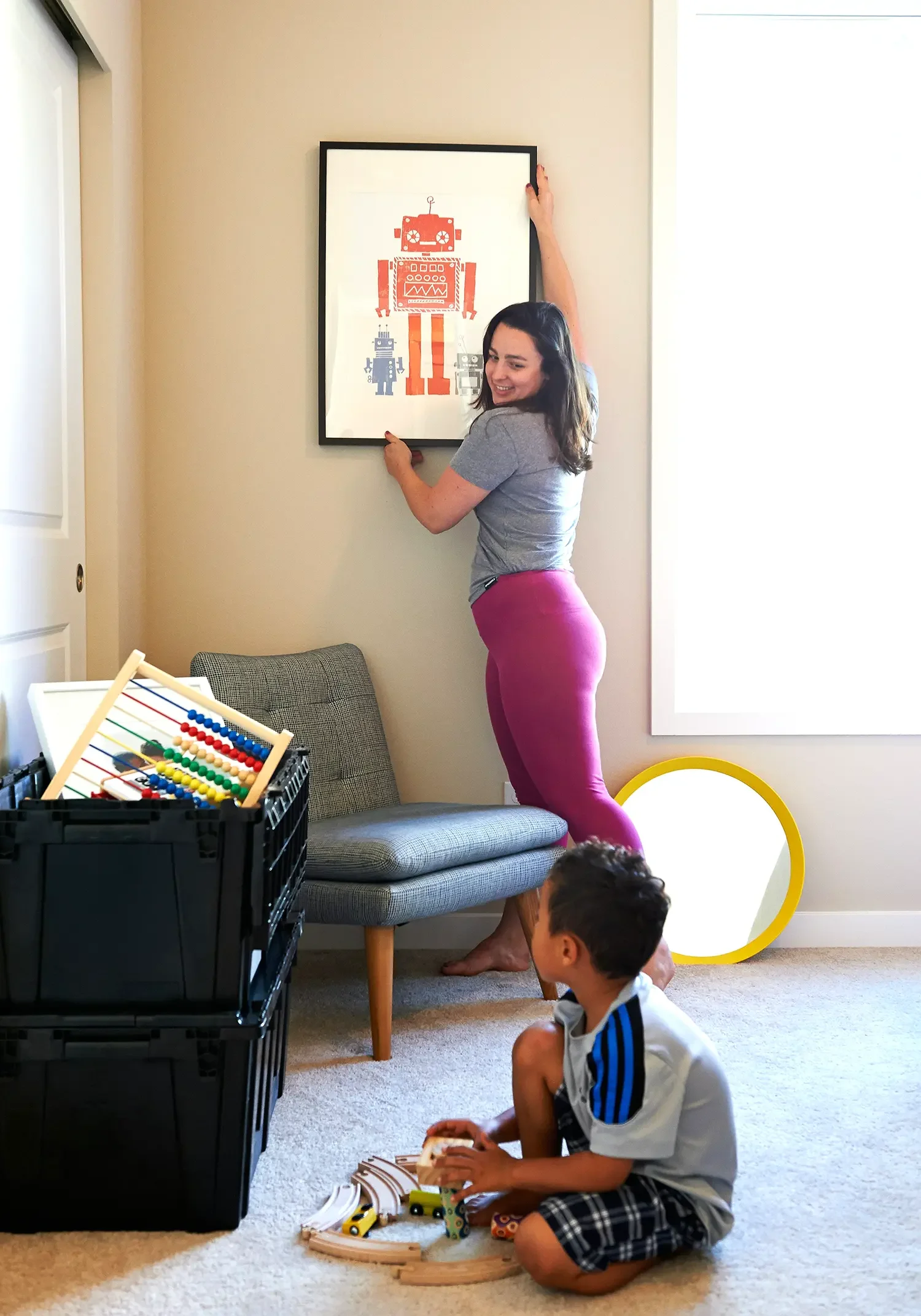 Una mujer coloca un cuadro con una ilustración de robots en una pared mientras que un niño juega con un tren de madera en el piso.