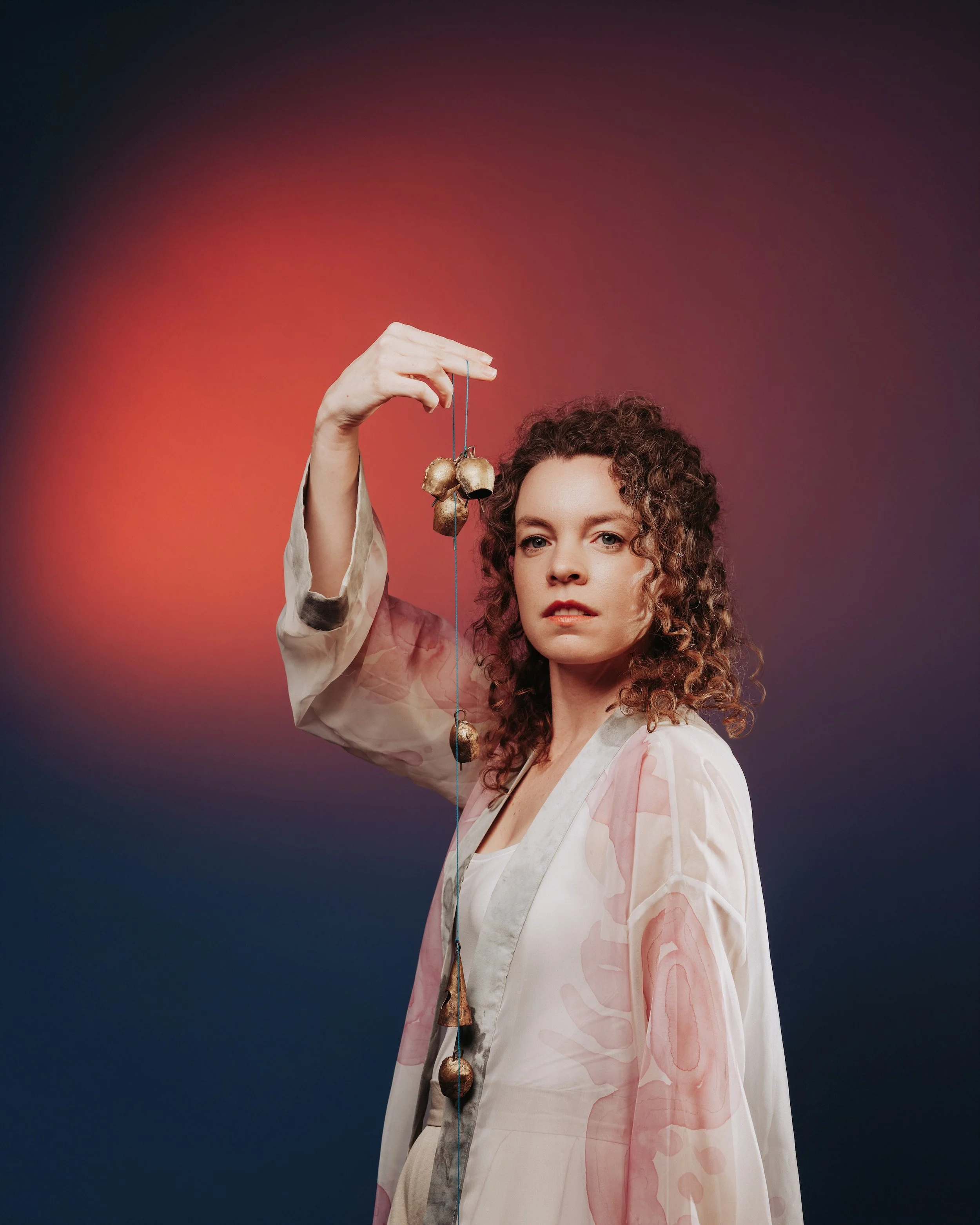 A photo of woman with curly hair wearing a white blouse, holding up a string of vintage bronze bells, standing against a dark red and purple gradient background.