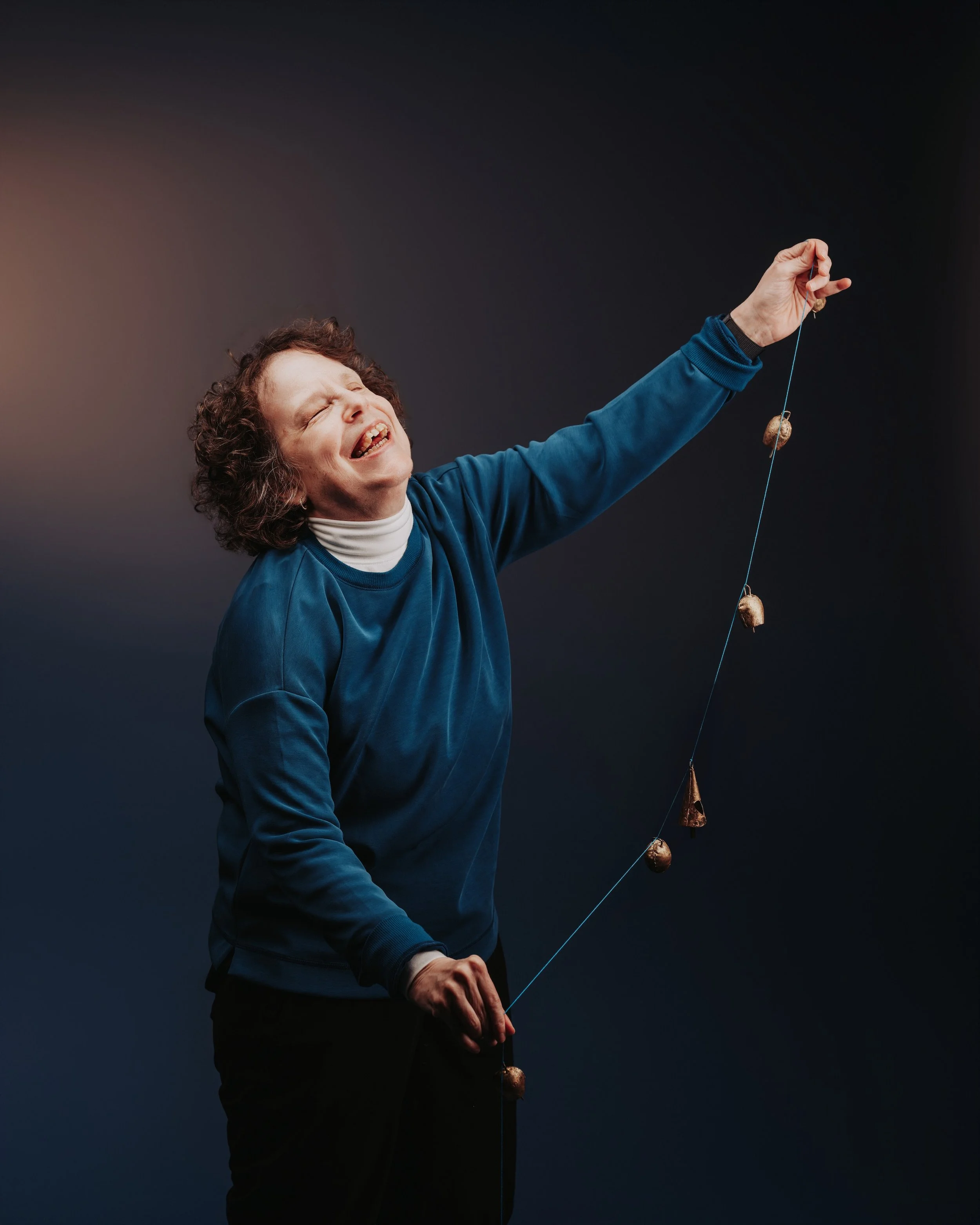 A photo of a smiling woman with curly hair wearing a blue sweater, holding up a blue string of vintage bronze bells, standing against a dark blue gradient background.