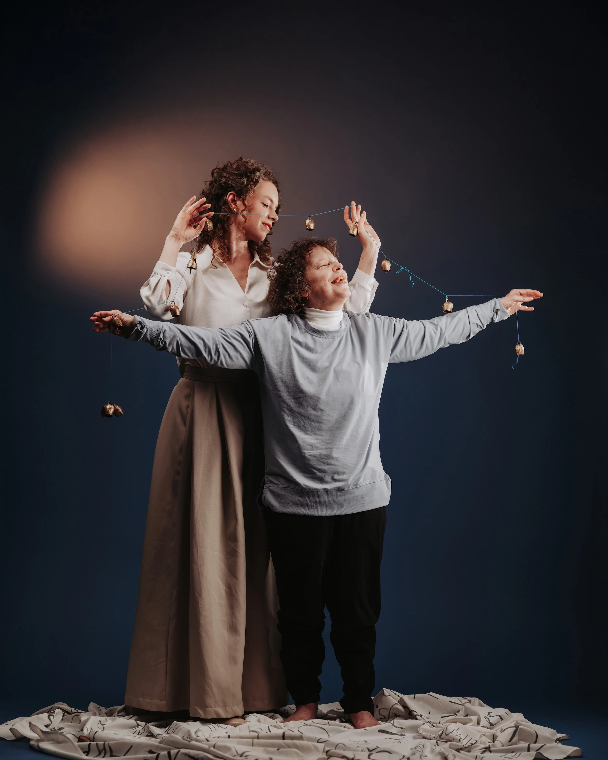 A photo of two women holding a bright blue string of vintage bronze bells against a dark blue backdrop. One stands behind in a white blouse with both arms raised, while the woman in grey stands in front with both arms stretched wide.