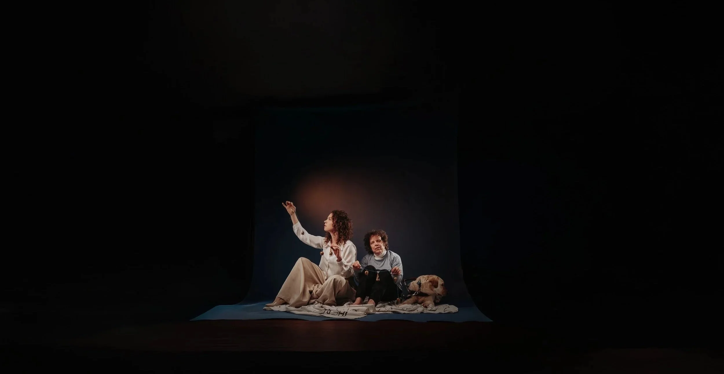 A photo of two women sitting on the floor against a dark background, holding hands and vintage bronze bells. A yellow labrador sits to the right of the older woman.
