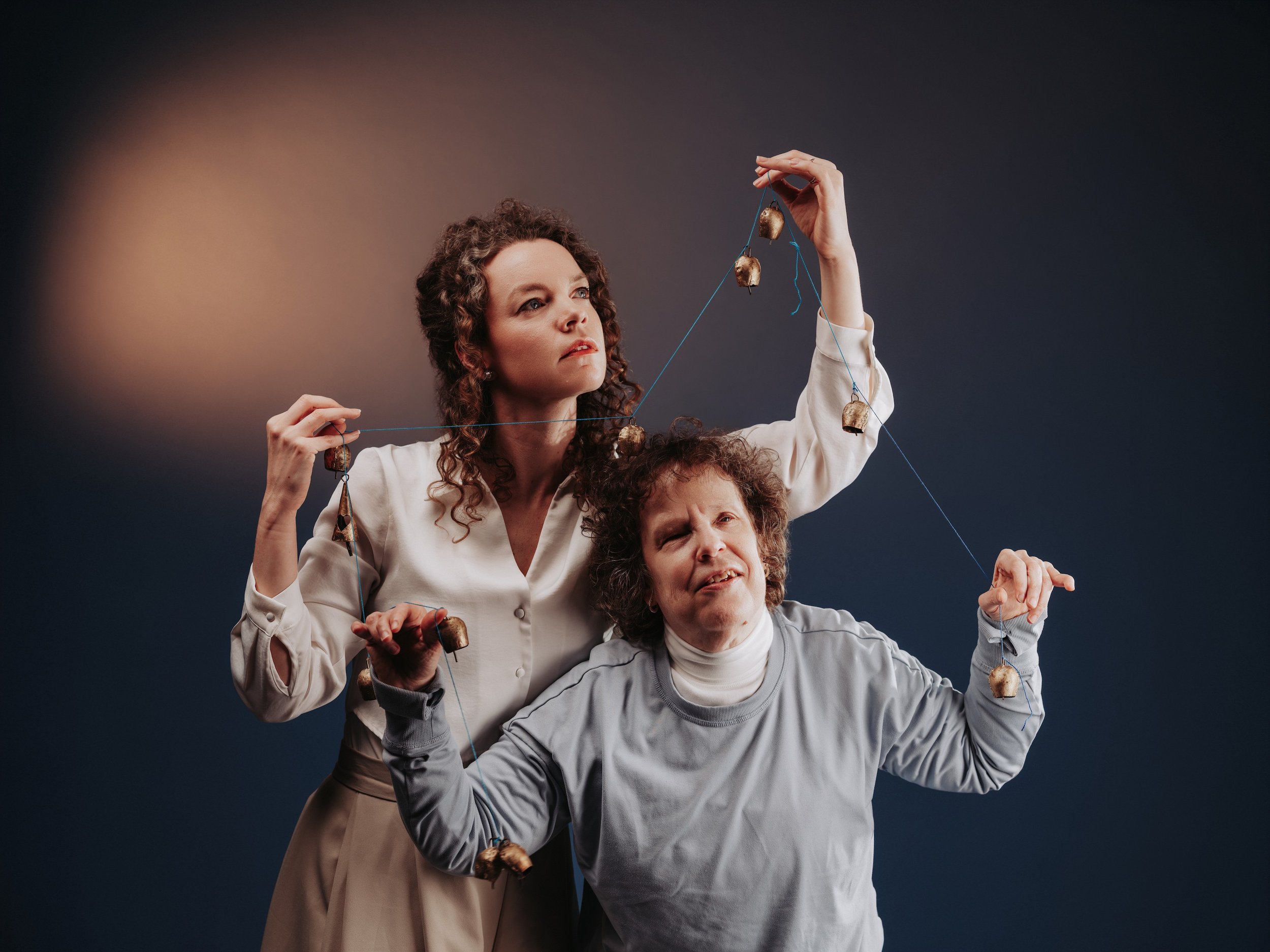A photo of two women posing against a dark blue backdrop, holding a bright blue string of vintage bronze bells. One stands behind in a white blouse with her arm raised, while the other stands in front wearing grey.