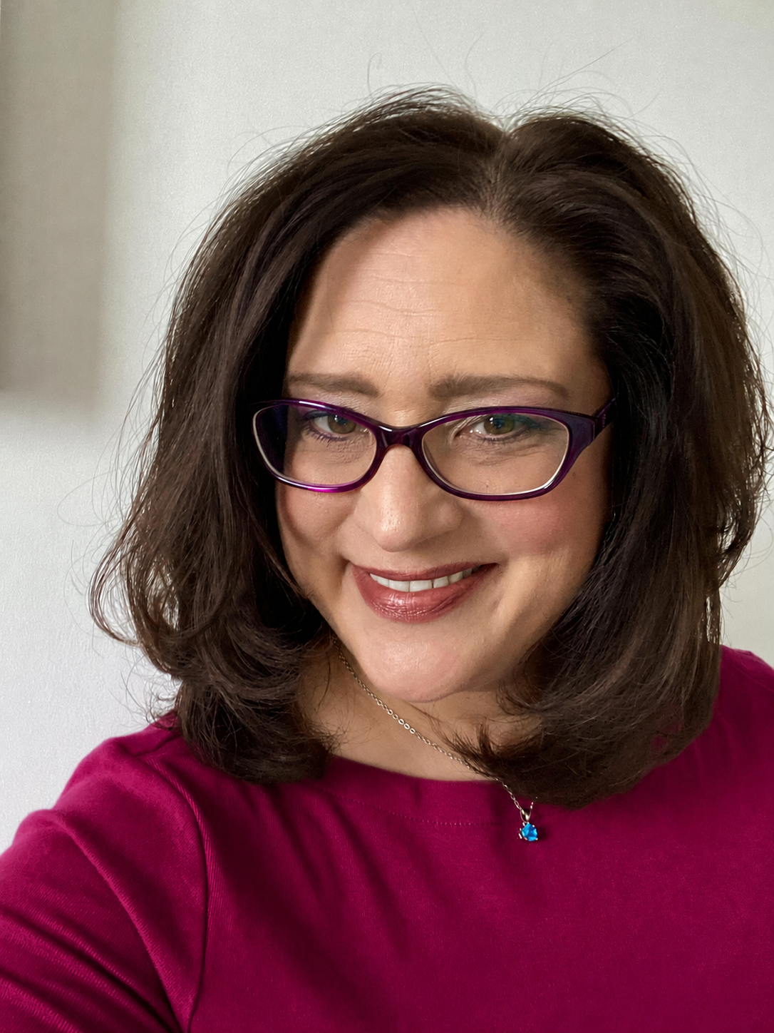 A woman with brown hair, wearing purple glasses and a magenta top, smiling, with a white background.