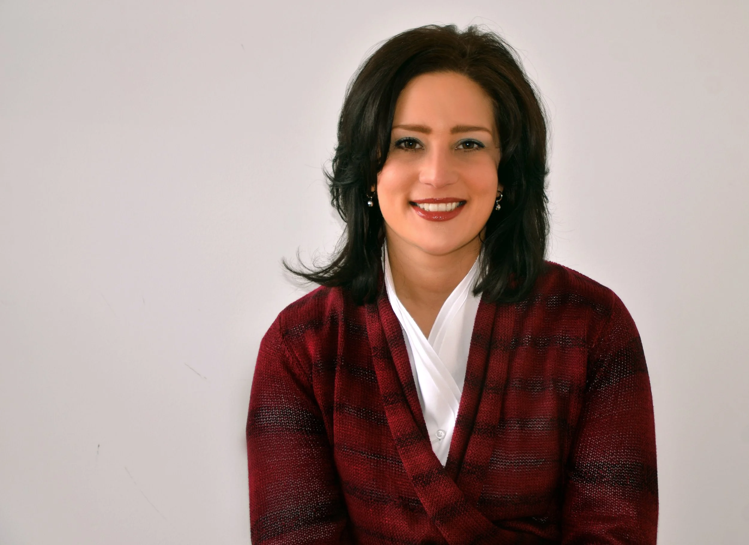 A woman with shoulder-length dark hair smiling, wearing a red and black checkered sweater over a white collared shirt, standing against a plain white background.