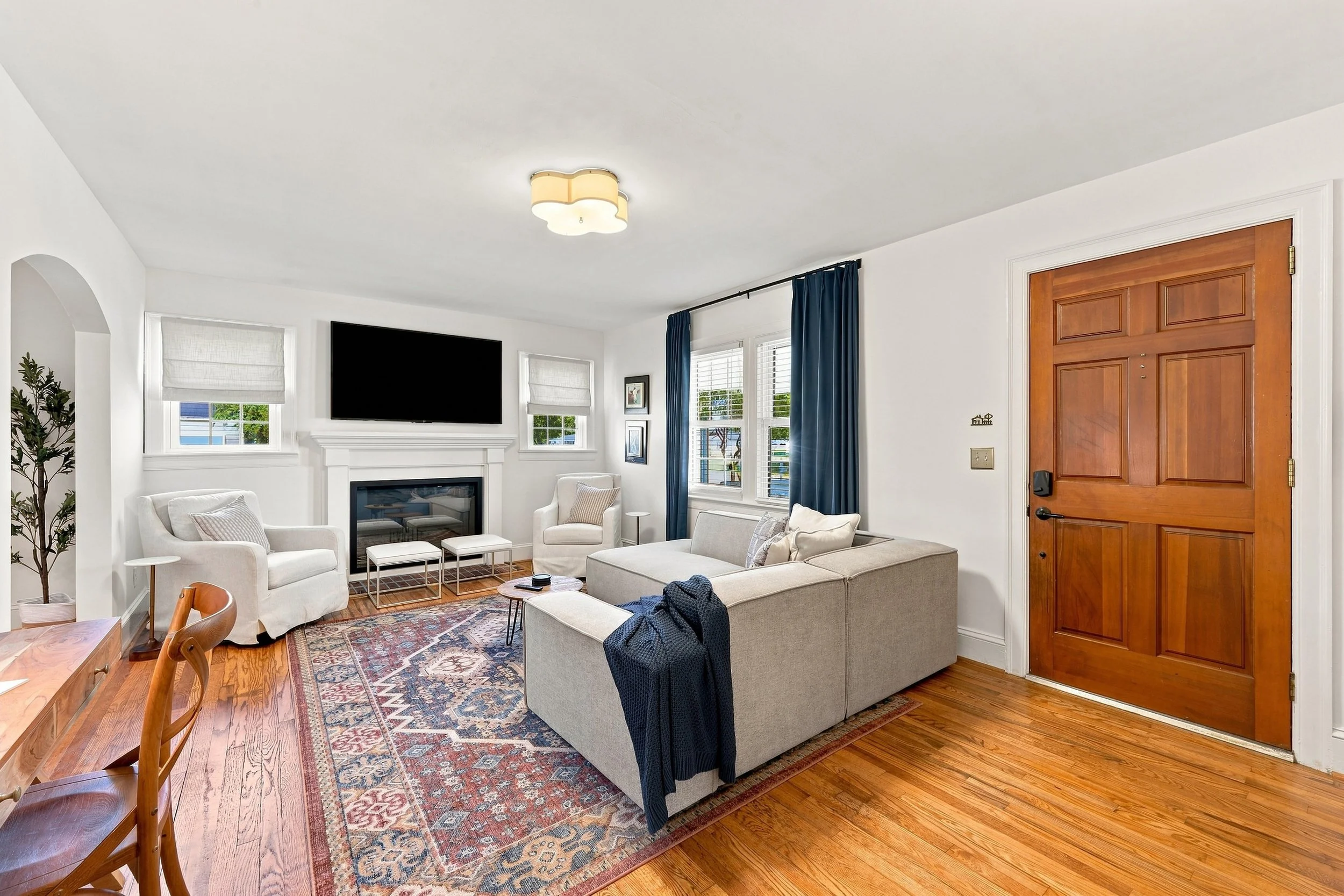 Living room with beige sofas, a patterned area rug, a fireplace with a flat-screen TV above it, two windows with white blinds, navy curtains, a wooden front door, and hardwood flooring.