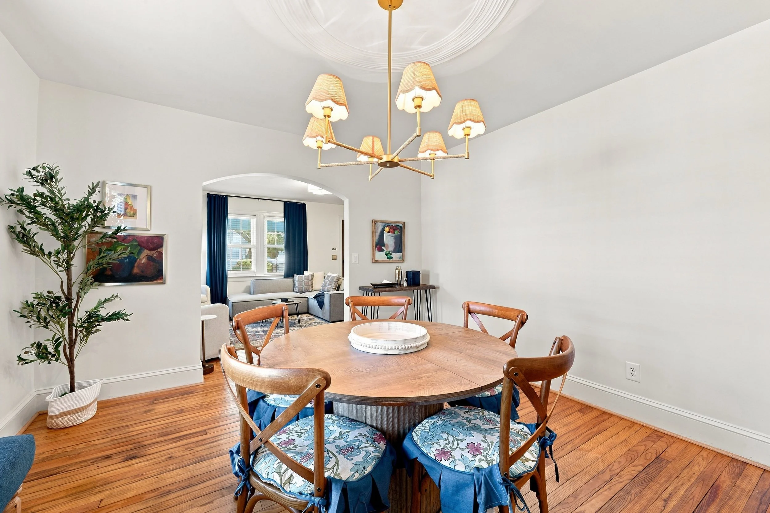 A dining room with a round wooden table surrounded by six chairs with floral-patterned cushions. A white decorative tray is on the table. The room has hardwood floors, white walls, and a large green plant in a white basket. In the background, there's