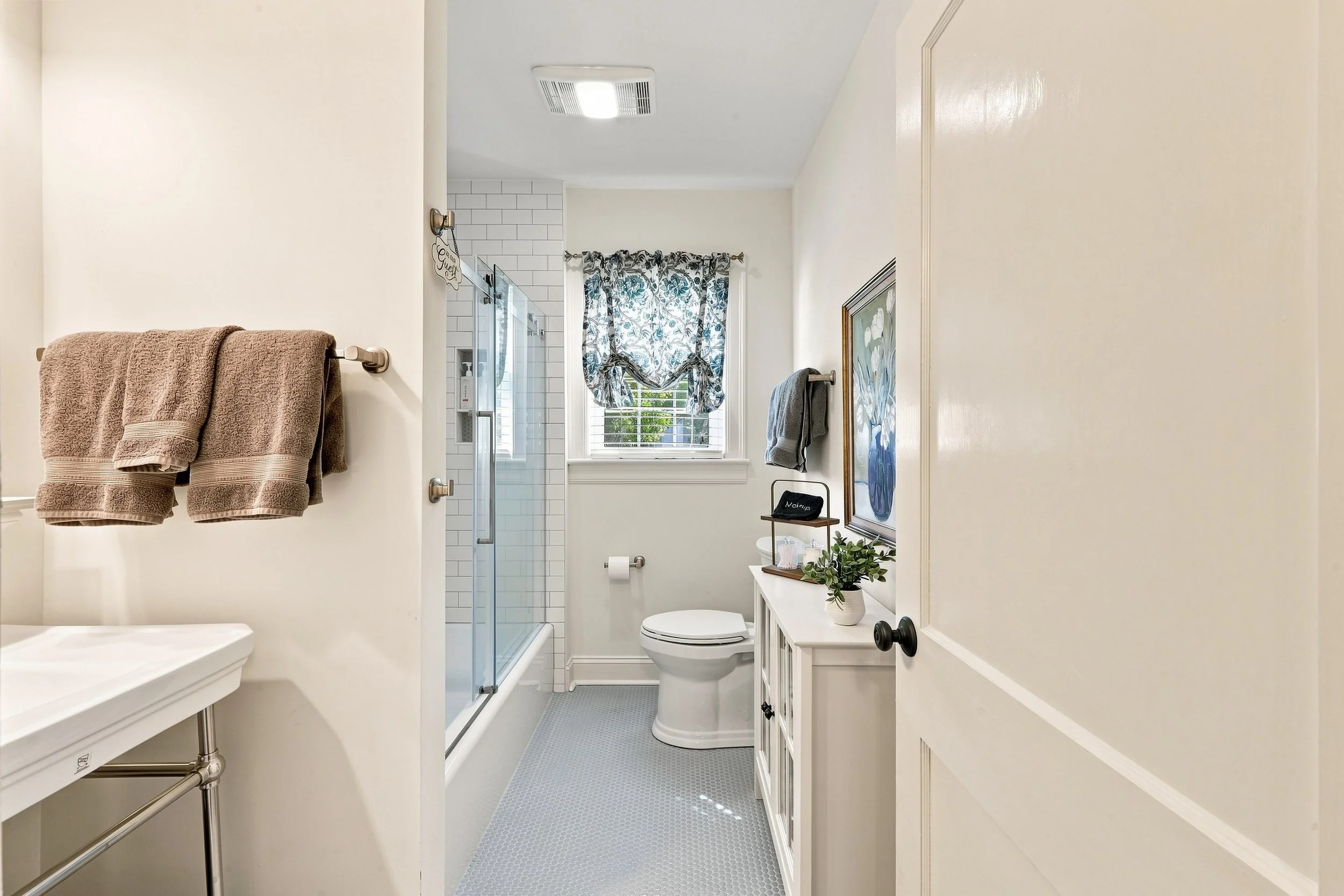 A bathroom interior with a white vanity on the left, two brown towels on a bar, a toilet below a window with floral curtains, a small shelf with a potted plant, and a shower with glass door in the background.