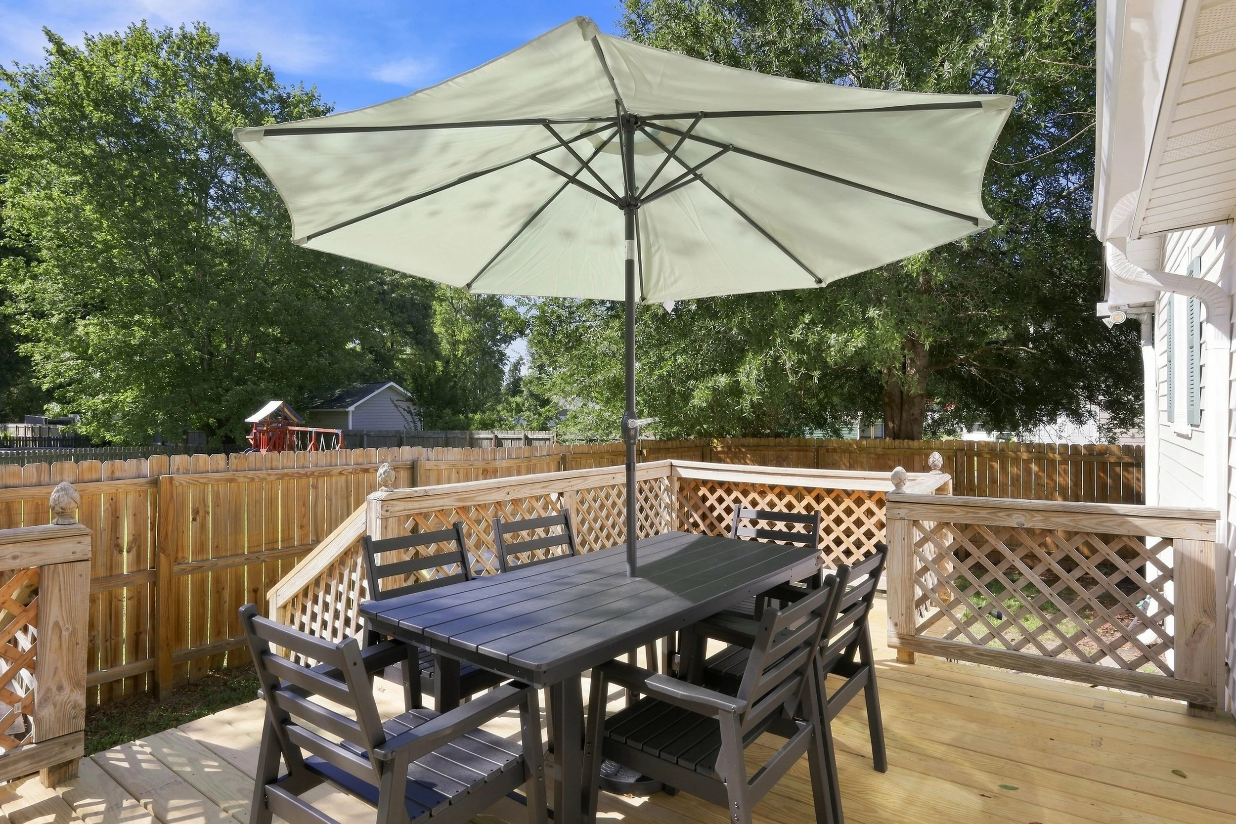 Backyard patio with black dining table and chairs, cream-colored umbrella, wooden fence, green trees, and blue sky.