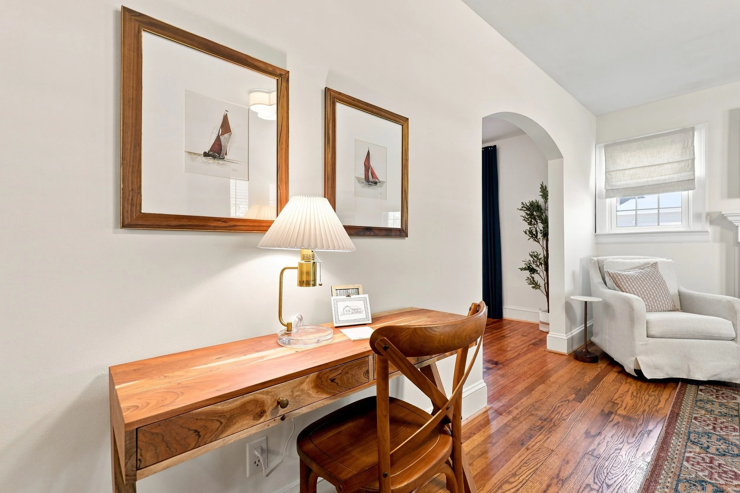 A cozy living room with white walls, wooden floors, and colorful rugs. There are two framed pictures of sailboats on the wall, a desk with a table lamp, a small plant, and a framed sketch. A white sofa with a pillow is visible in the corner near a wi