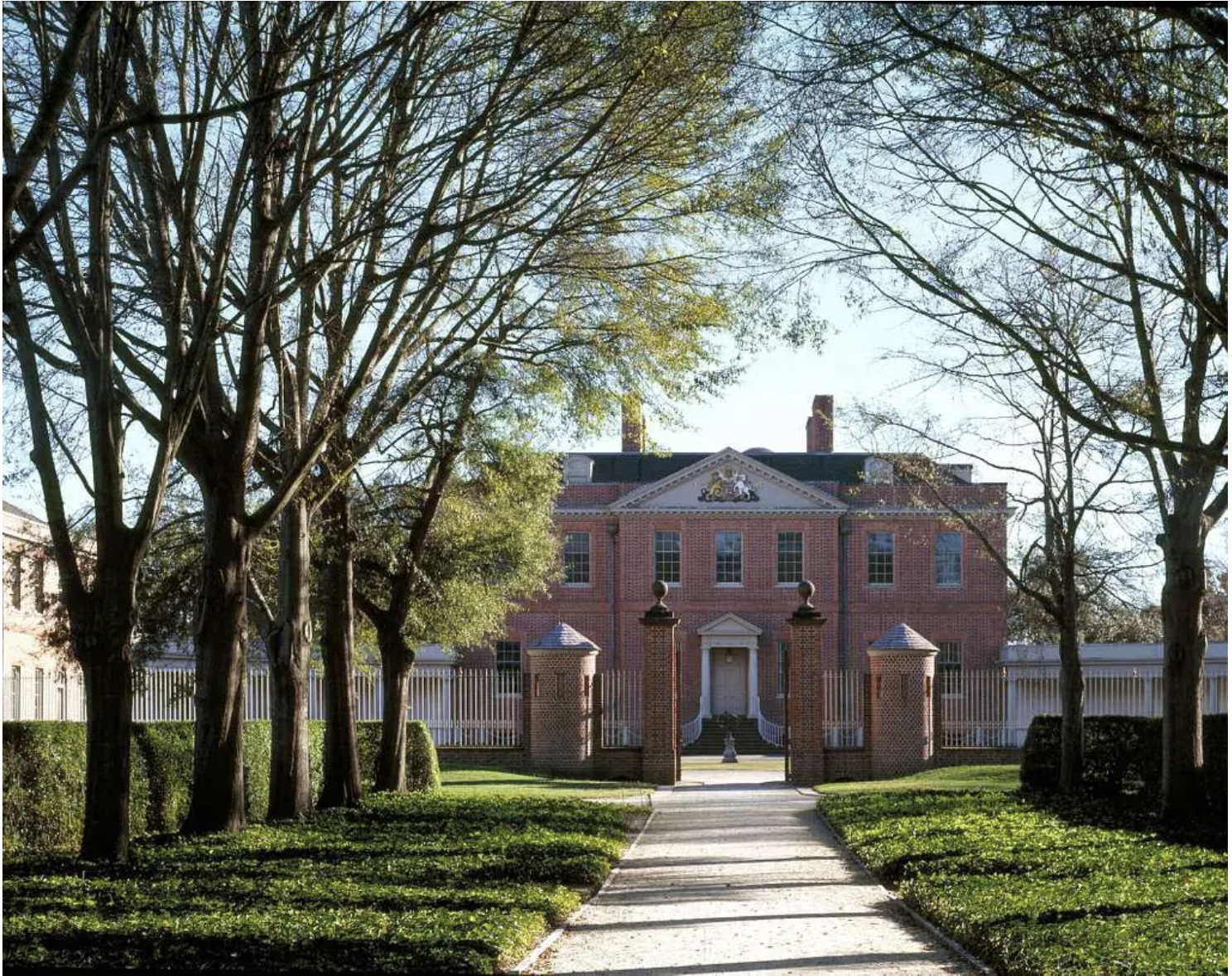 A large brick mansion with a white door and a triangular pediment, surrounded by tall trees and a white picket fence, with a pathway leading to the entrance.