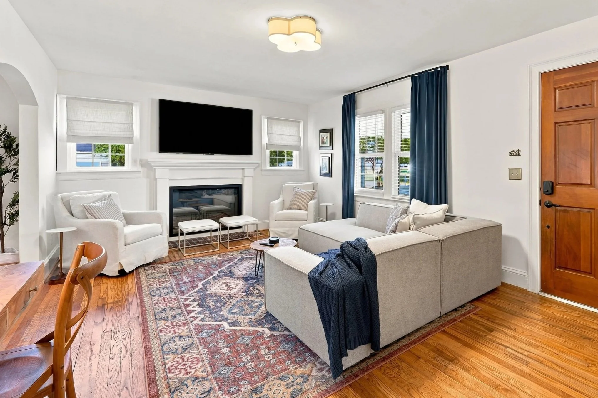 Living room with white walls, hardwood floors, a light-colored sofa, a white armchair, a fireplace, a mounted flat-screen TV, windows with white blinds, blue curtains, framed artwork, and a patterned area rug.
