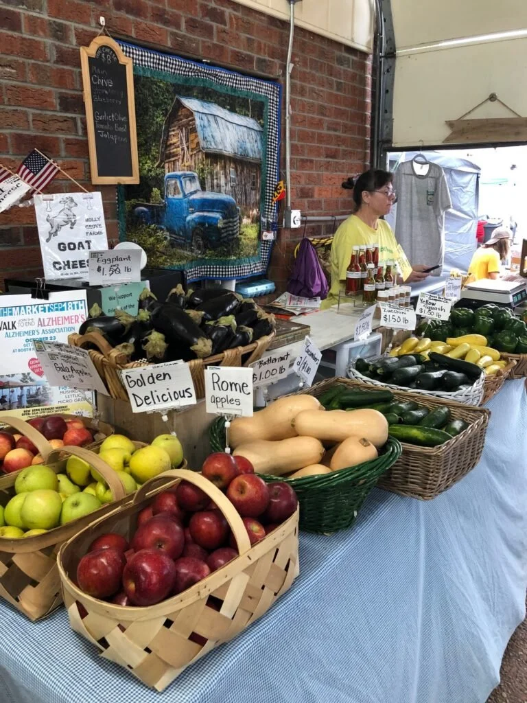 A farmers market stall displaying baskets of apples, zucchini, and butternut squash, with a woman selling produce, signs, and a painted rustic barn scene in the background.