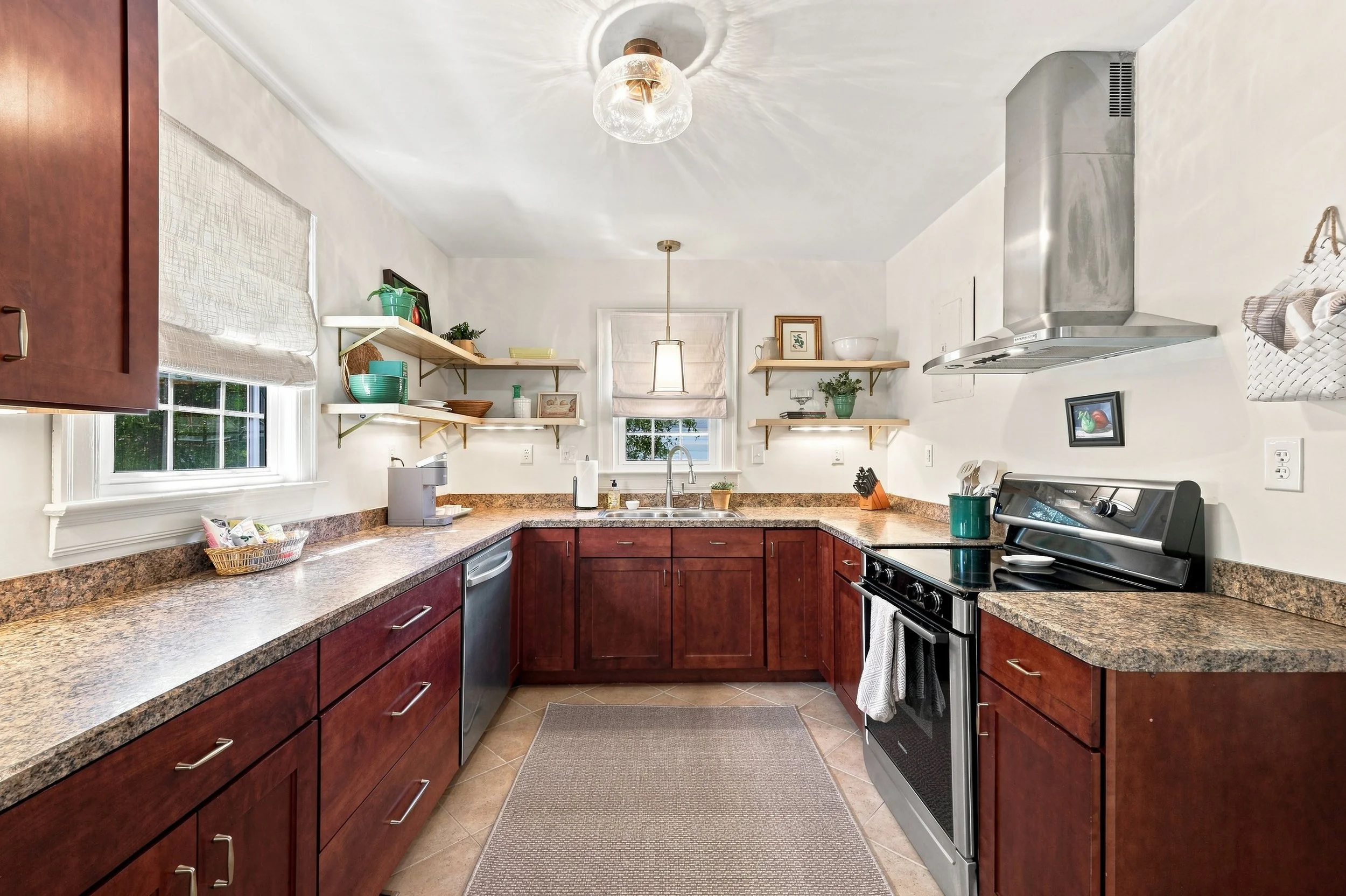 A kitchen with granite countertops, wooden cabinets, stainless steel appliances including an oven, and open shelves with decorative items. There are two windows with white Roman shades and a ceiling light and pendant light. The floor is tiled, and th