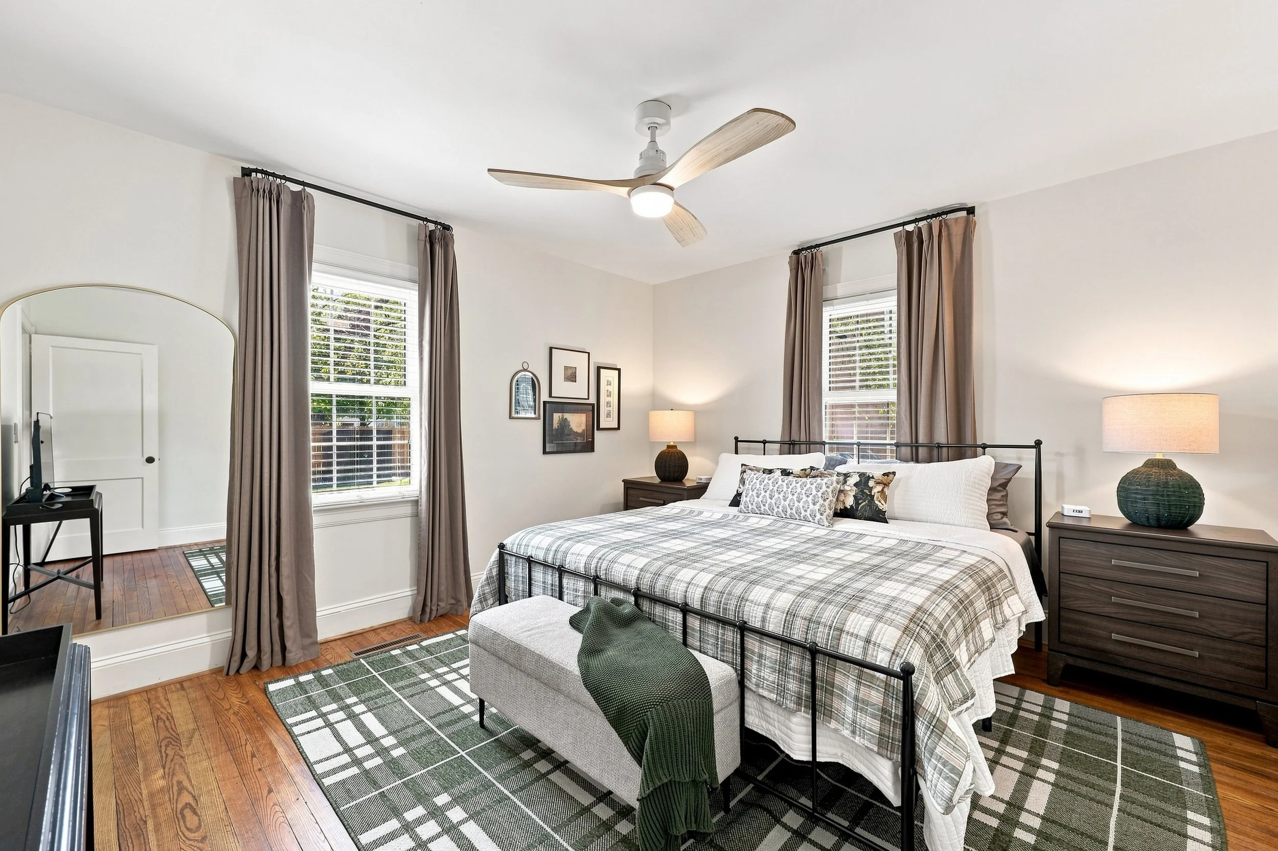 A bedroom with a bed, two windows with curtains, a ceiling fan, a mirror, a dresser, and a nightstand with a lamp, decorated in neutral tones with plants visible outside the windows.
