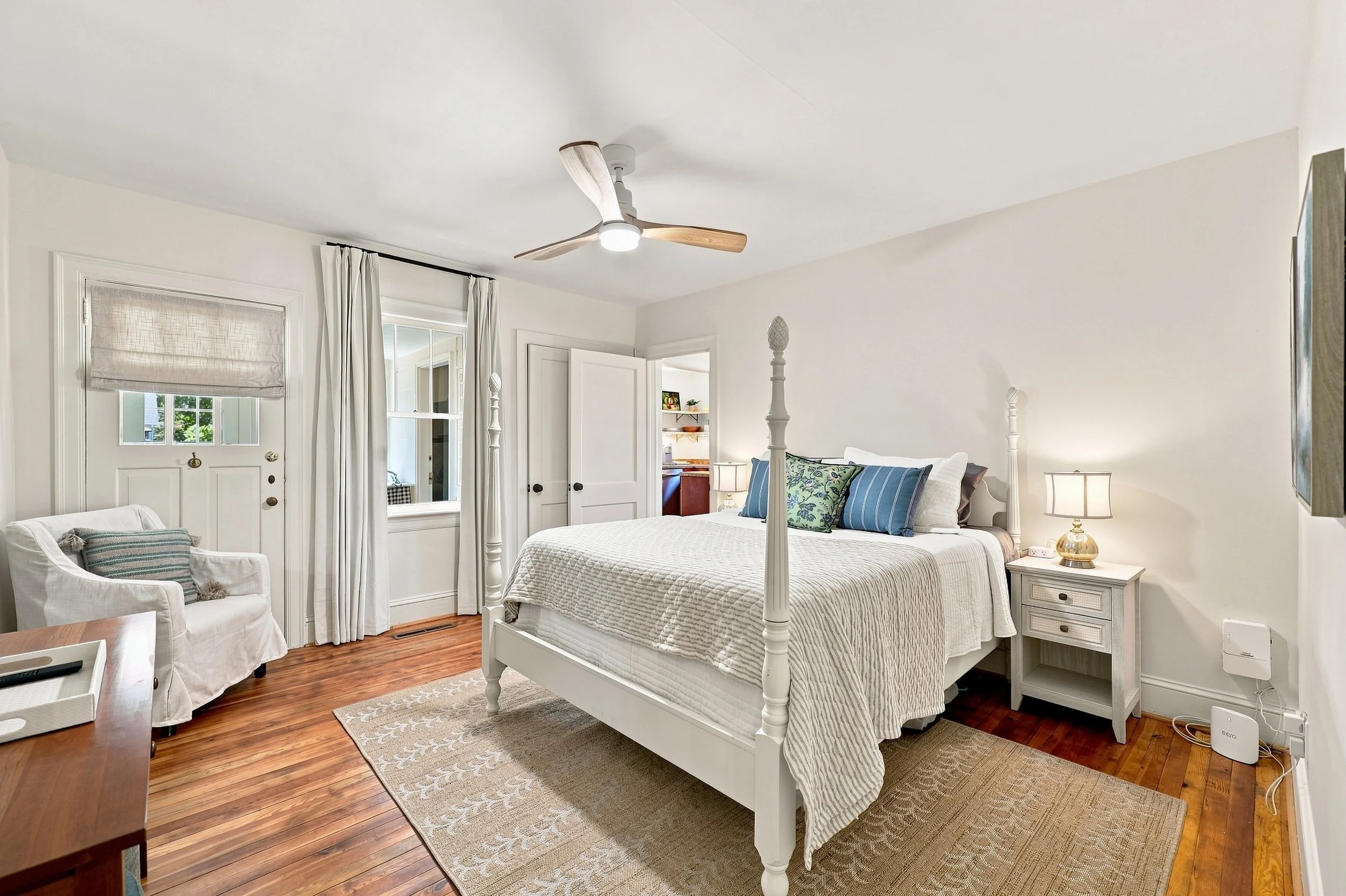 A bedroom with a white four-poster bed, a nightstand with a lamp, an armchair with pillows, a window with curtains, hardwood floors, and a ceiling fan.