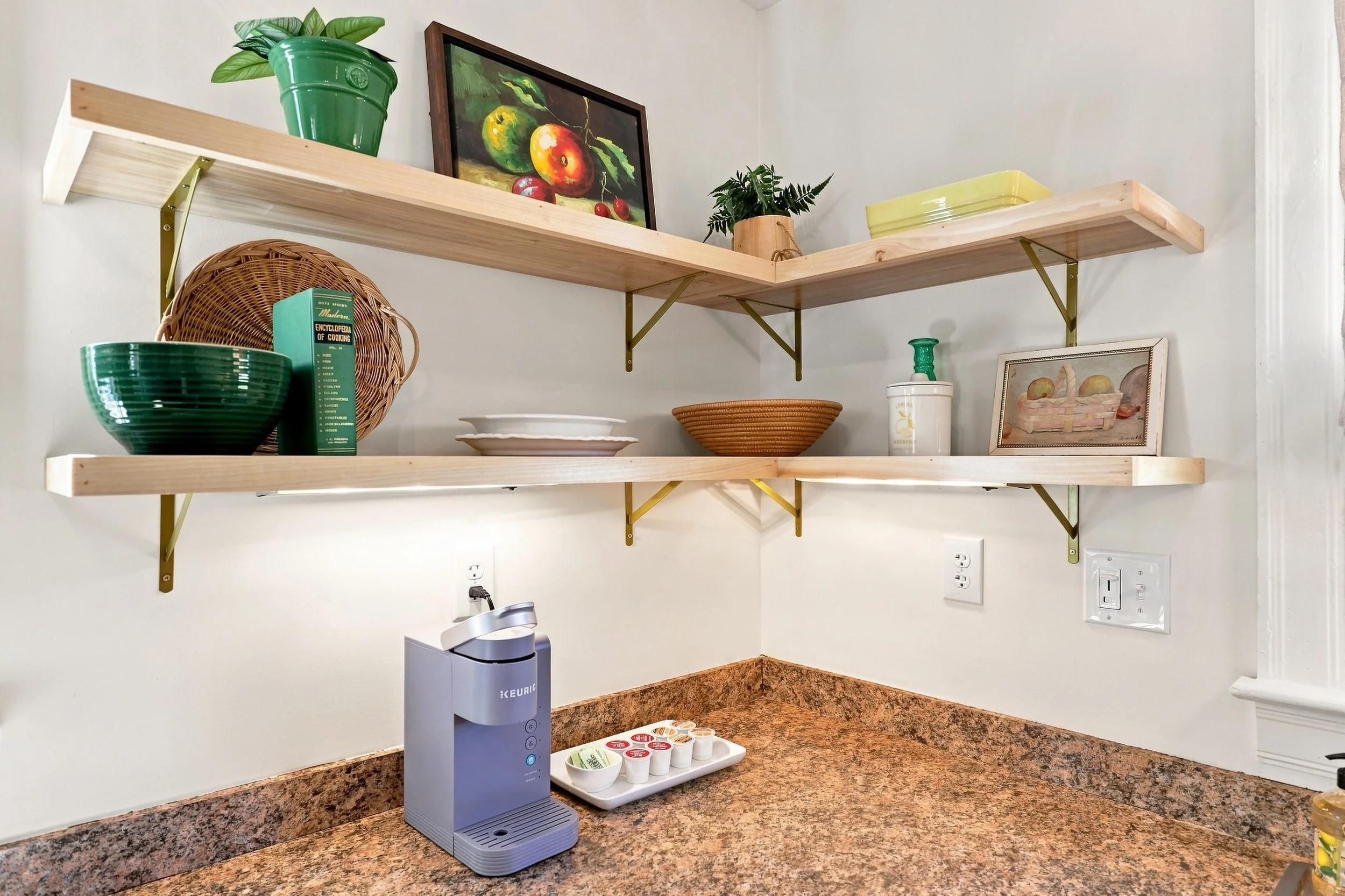 Kitchen corner with two wooden shelves holding a green plant in a green pot, a framed painting of apples, a yellow container, and a small framed picture of a basket of fruit. The countertop below has a blue Keurig coffee maker, a tray with coffee pod