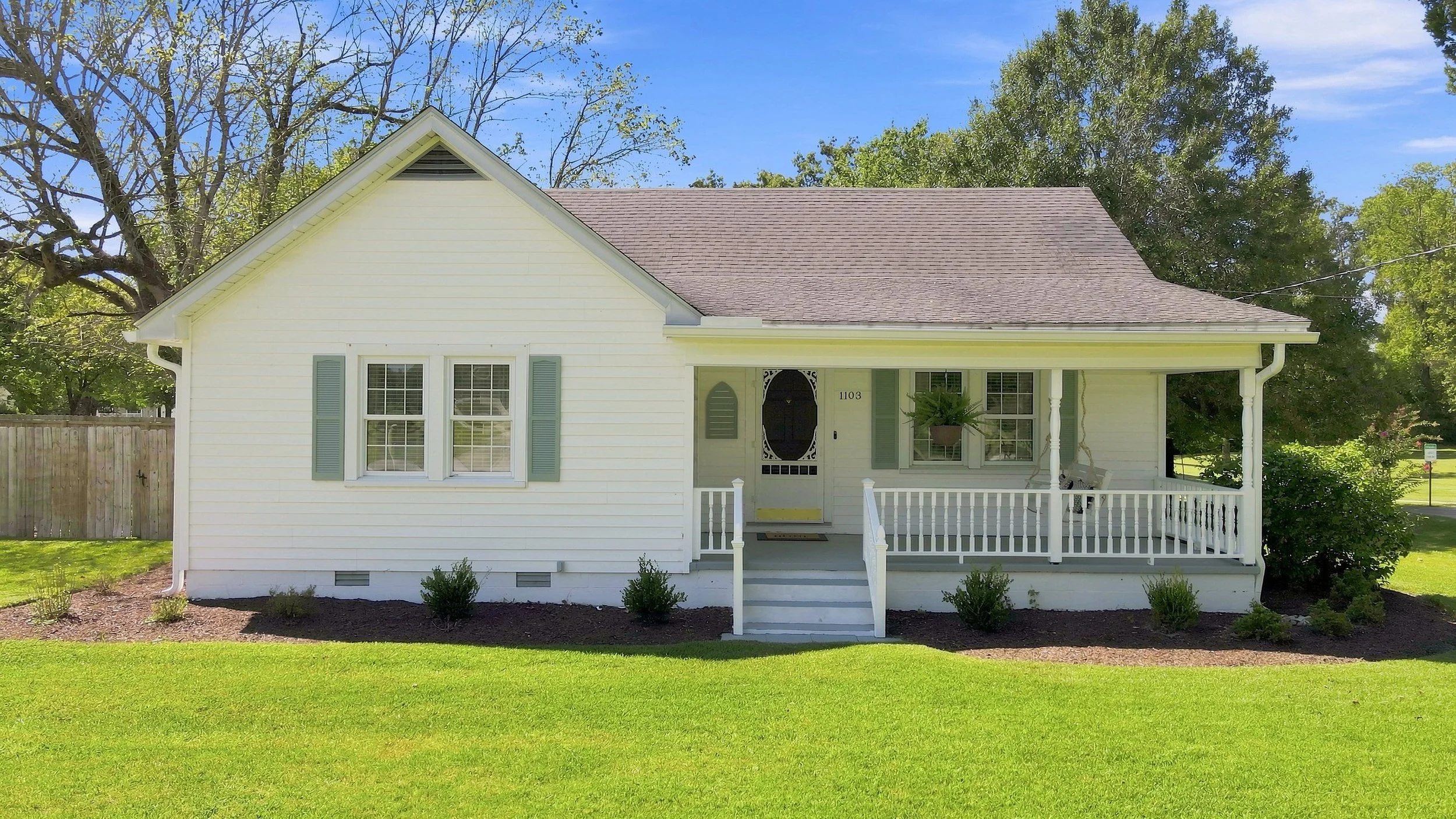 A white house with a front porch, light blue shutters, and a brown roof, surrounded by green lawn and trees, under a clear blue sky.