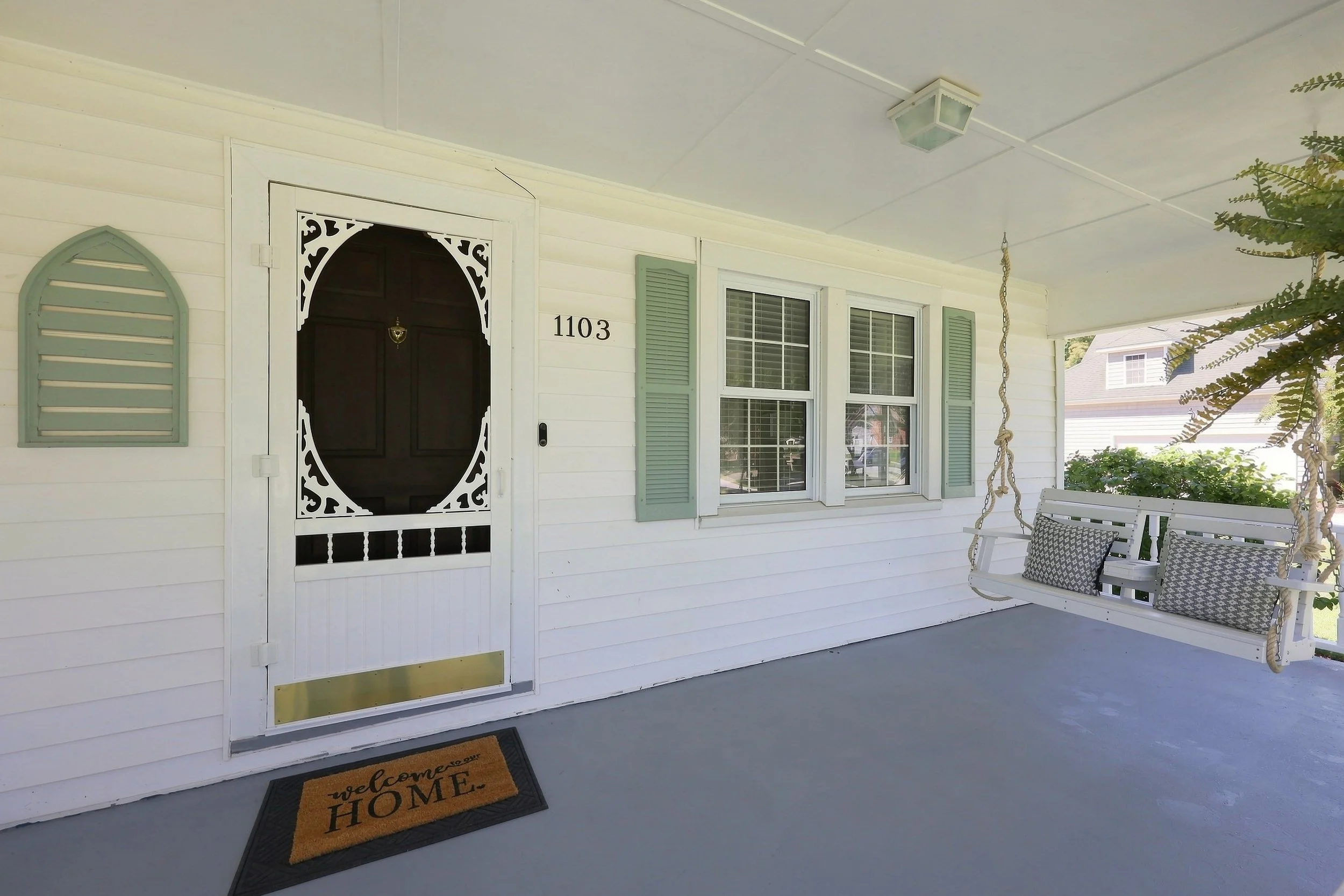 Front porch of a house with a black door, white siding, and light green shutters next to a window. A porch swing with cushions and a rope swing are present. There is a welcome mat on the floor with the words 'welcome HOME'.