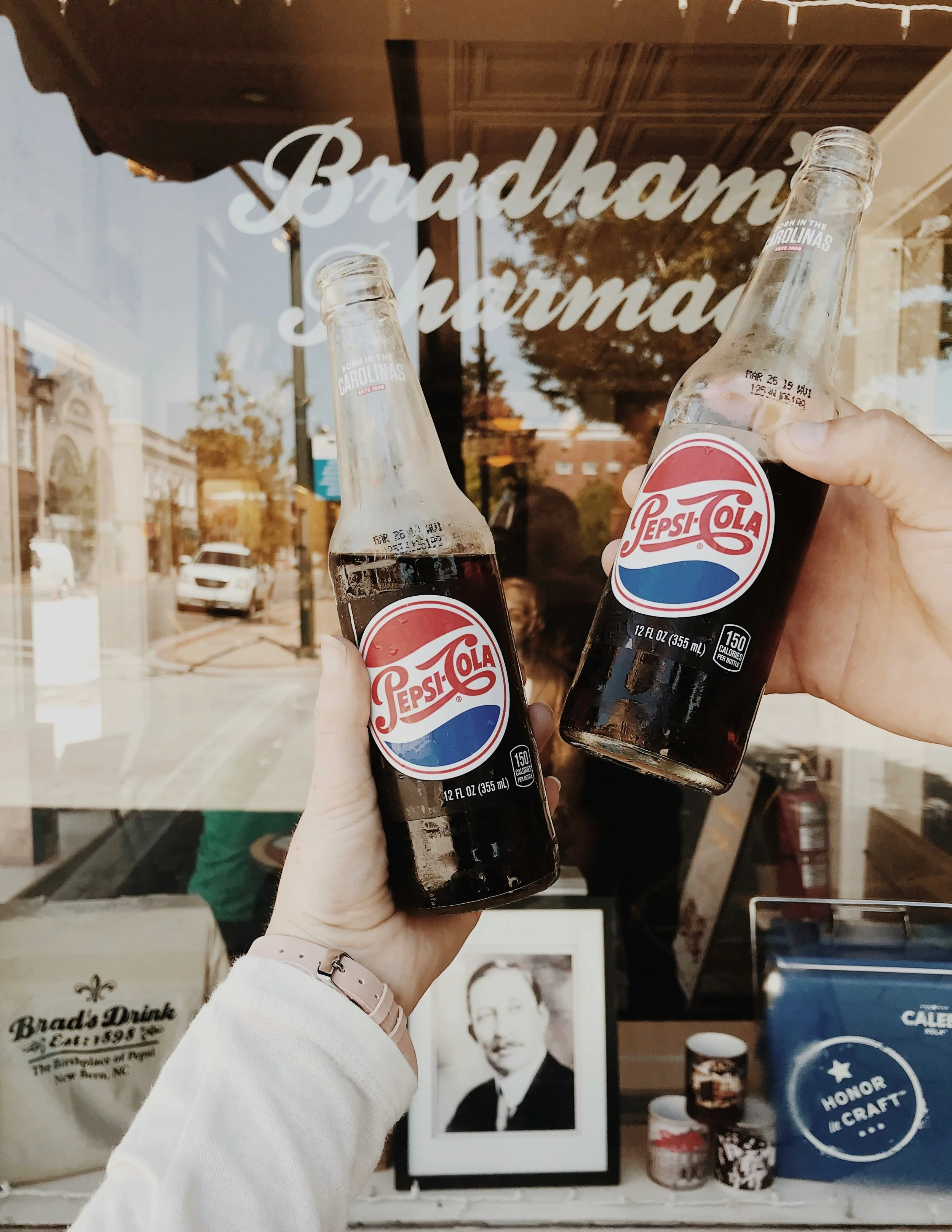 Two people holding glass bottles of Pepsi-Cola in front of a store window with the store name 'Bradham Pharm'. Reflection of the street and trees in the window, with signs and name tags visible inside.