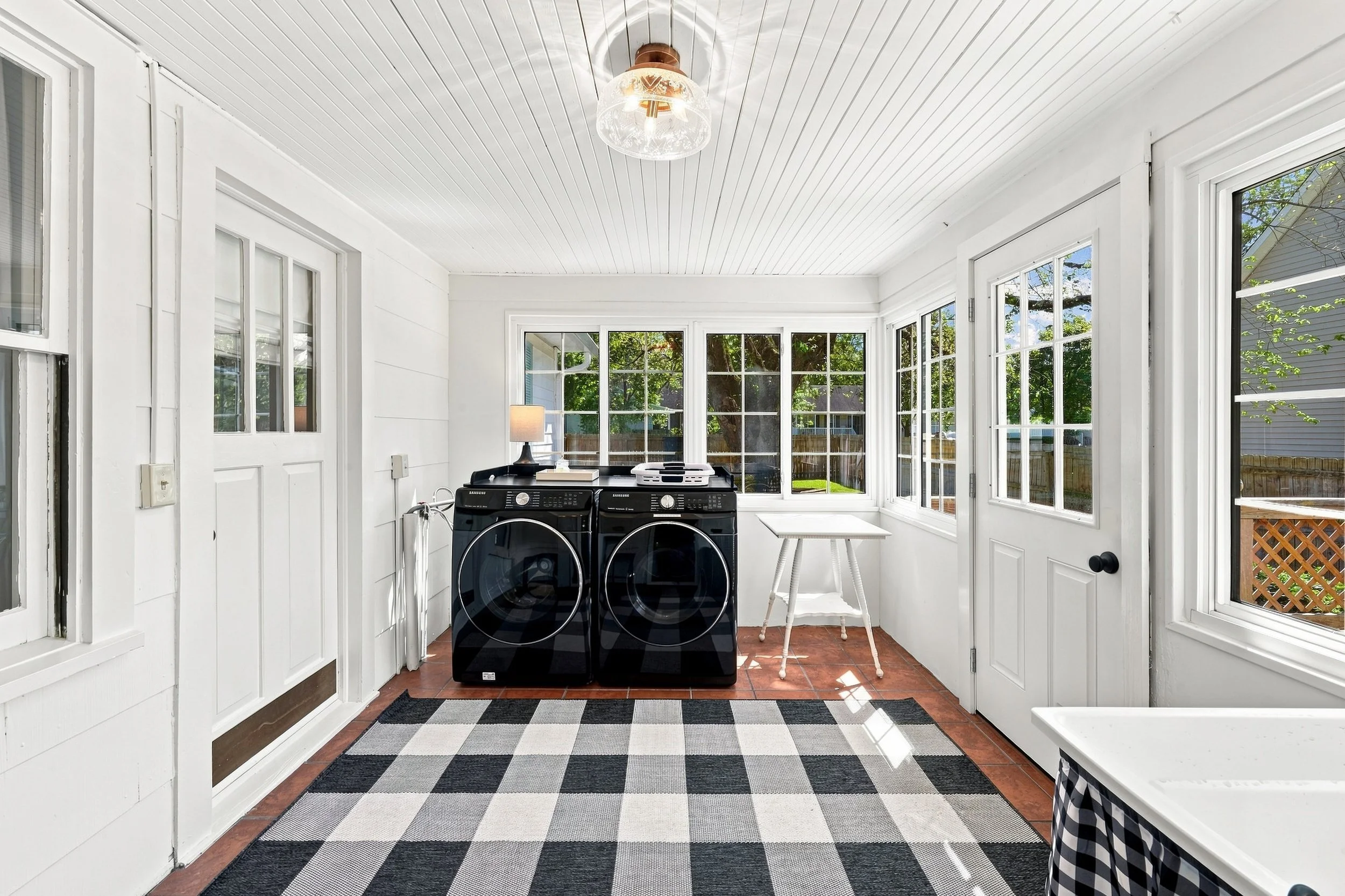 Sunroom with white walls, windows, laundry appliances, a small table, a lamp, and a checkered rug.