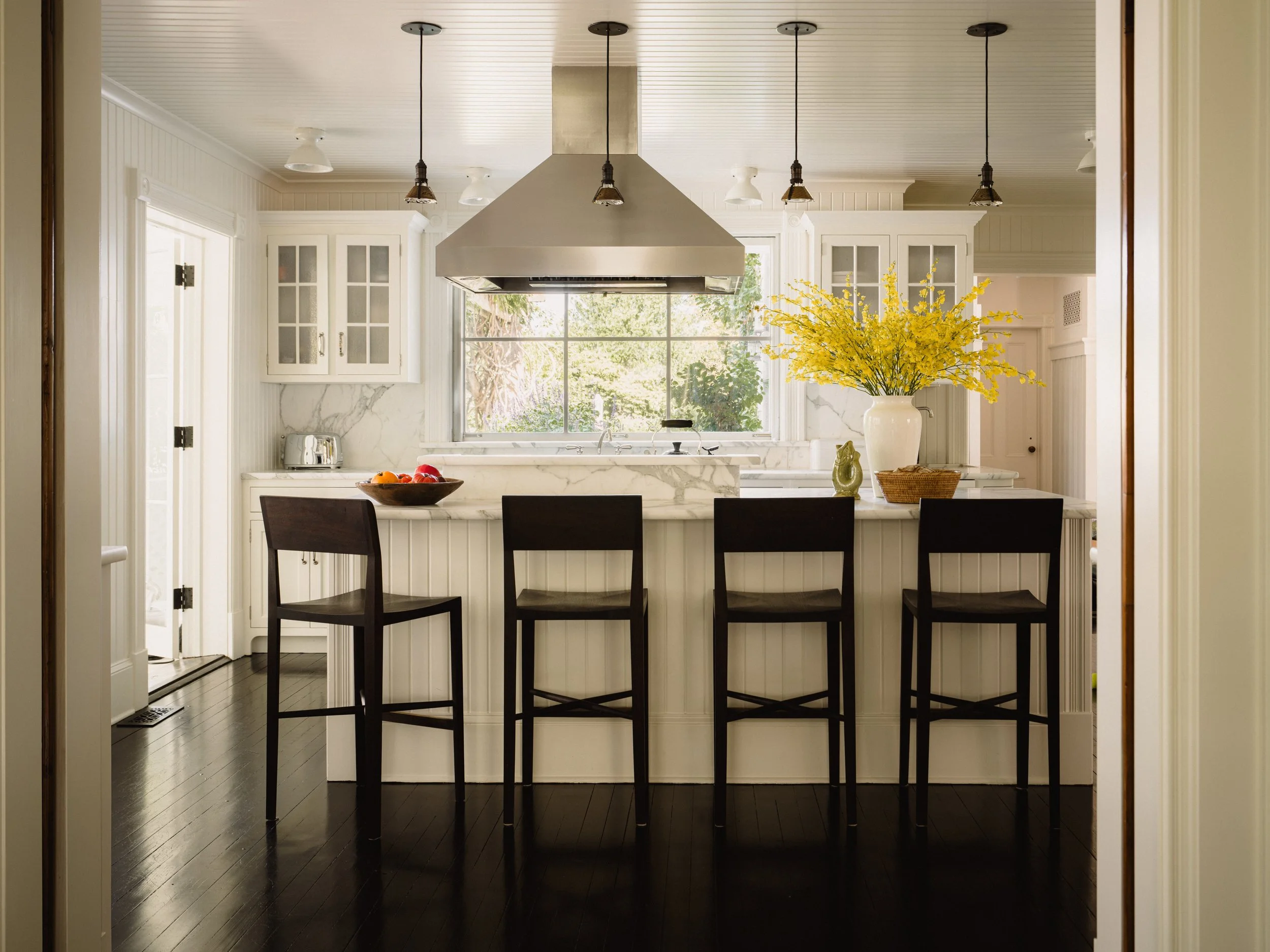 A modern kitchen with a large window, white cabinets with glass doors, a marble countertop, a yellow flower arrangement in a vase, a bowl of fruit, and three dark wooden barstools.